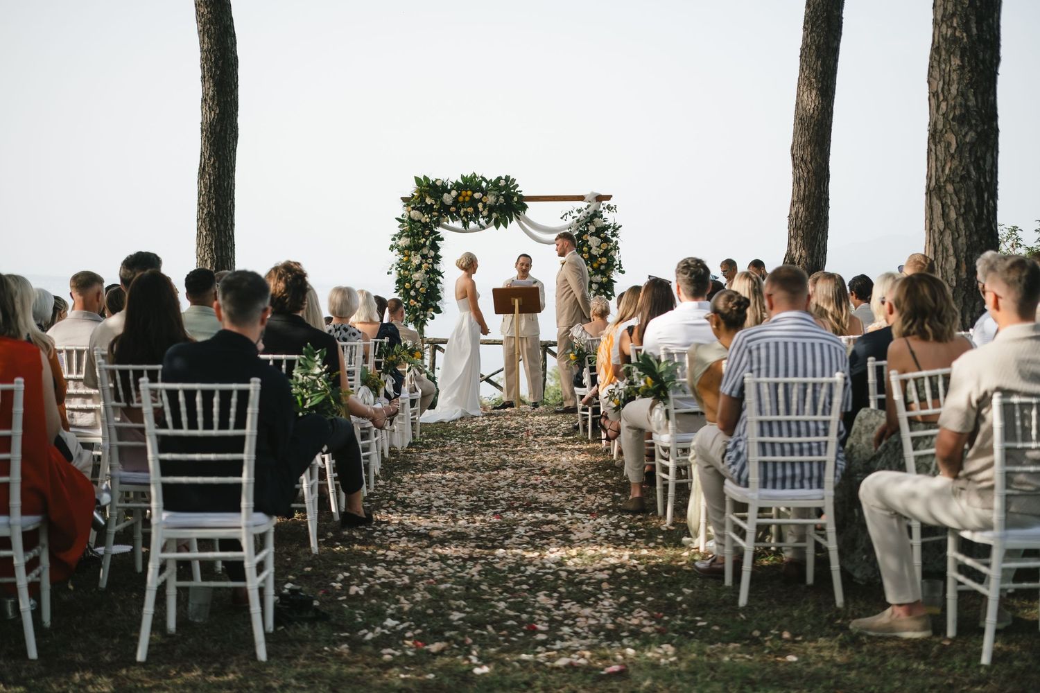 Outdoor wedding ceremony with guests seated along a petal-lined aisle under trees.