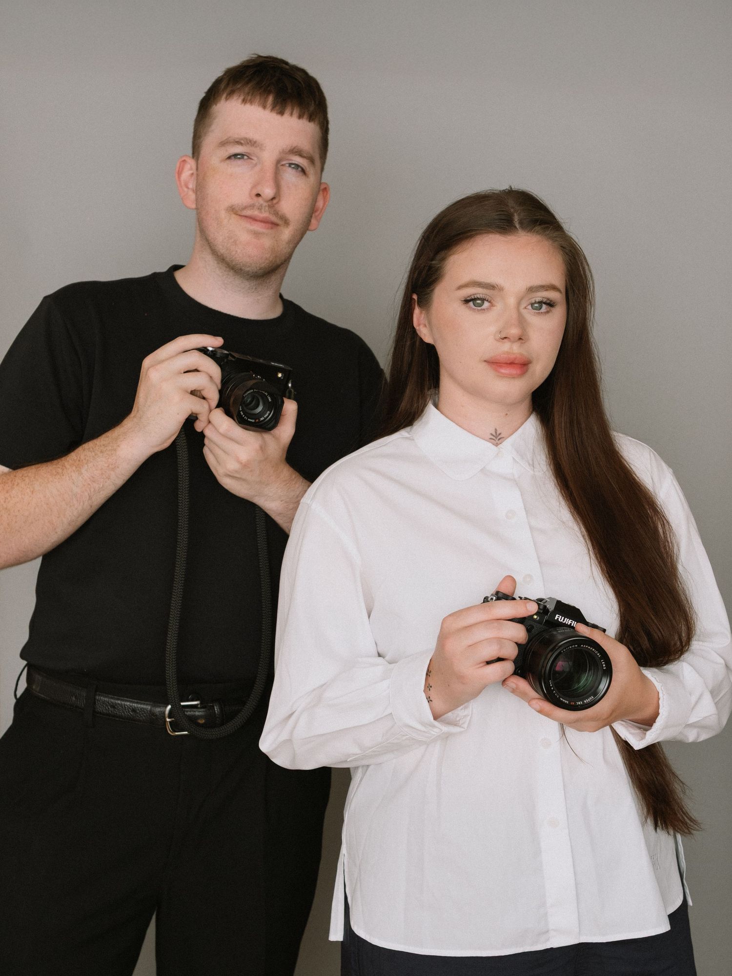 Two professional photographers holding cameras pose against a grey studio background.