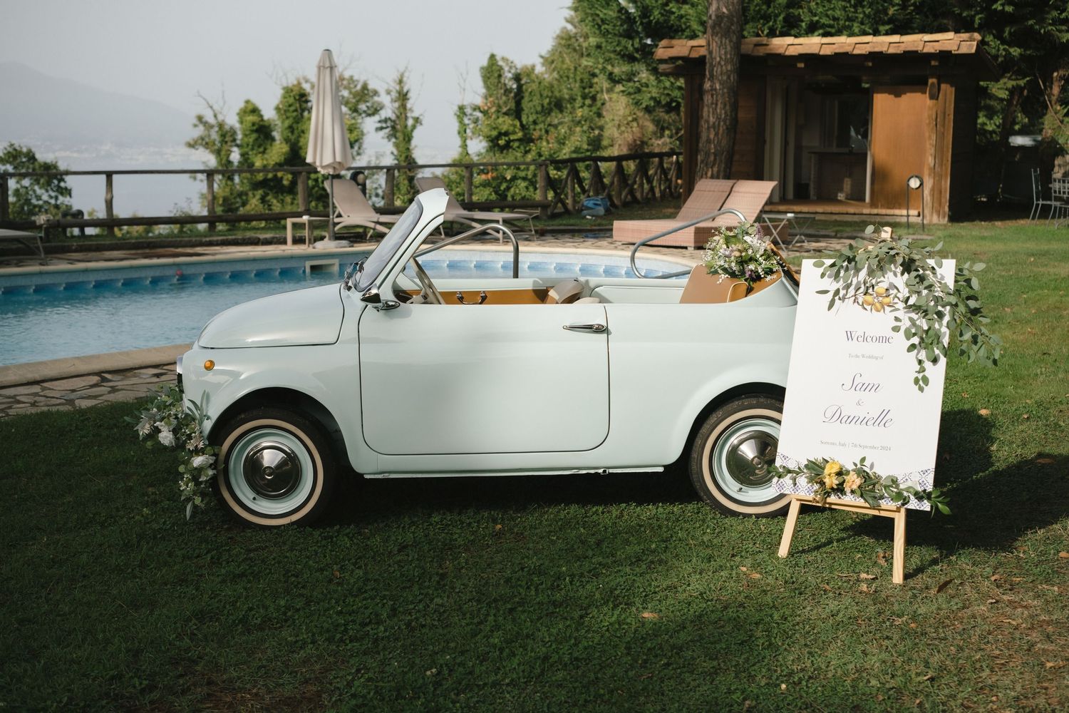 Vintage light blue Fiat 500 convertible decorated with flowers at a wedding venue beside a pool.