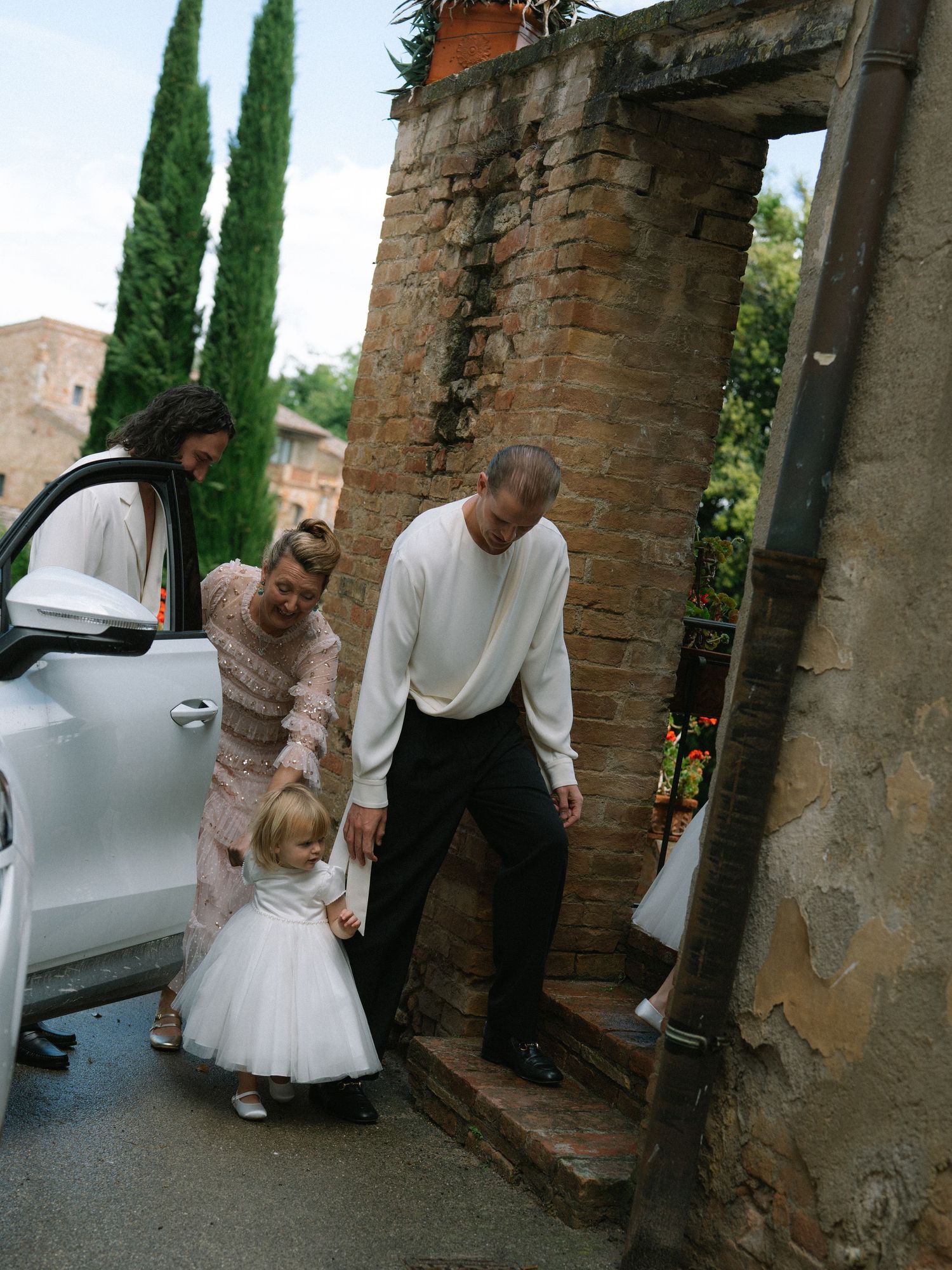 Family with toddler in white dress arriving at rustic Tuscan wedding venue by car.