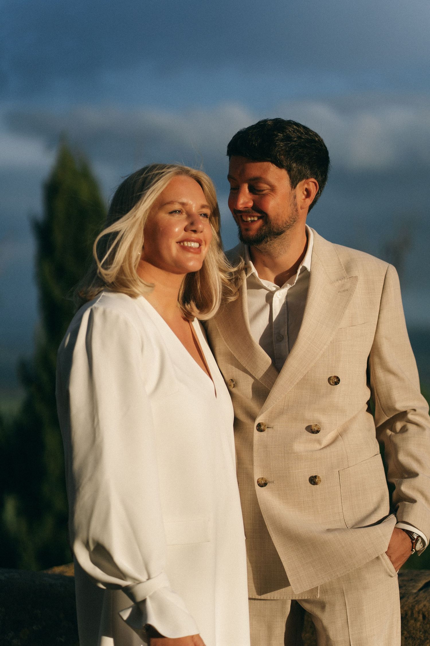A smiling blonde bride in white blouse stands with her groom in a beige suit against a dramatic Tuscan sky.