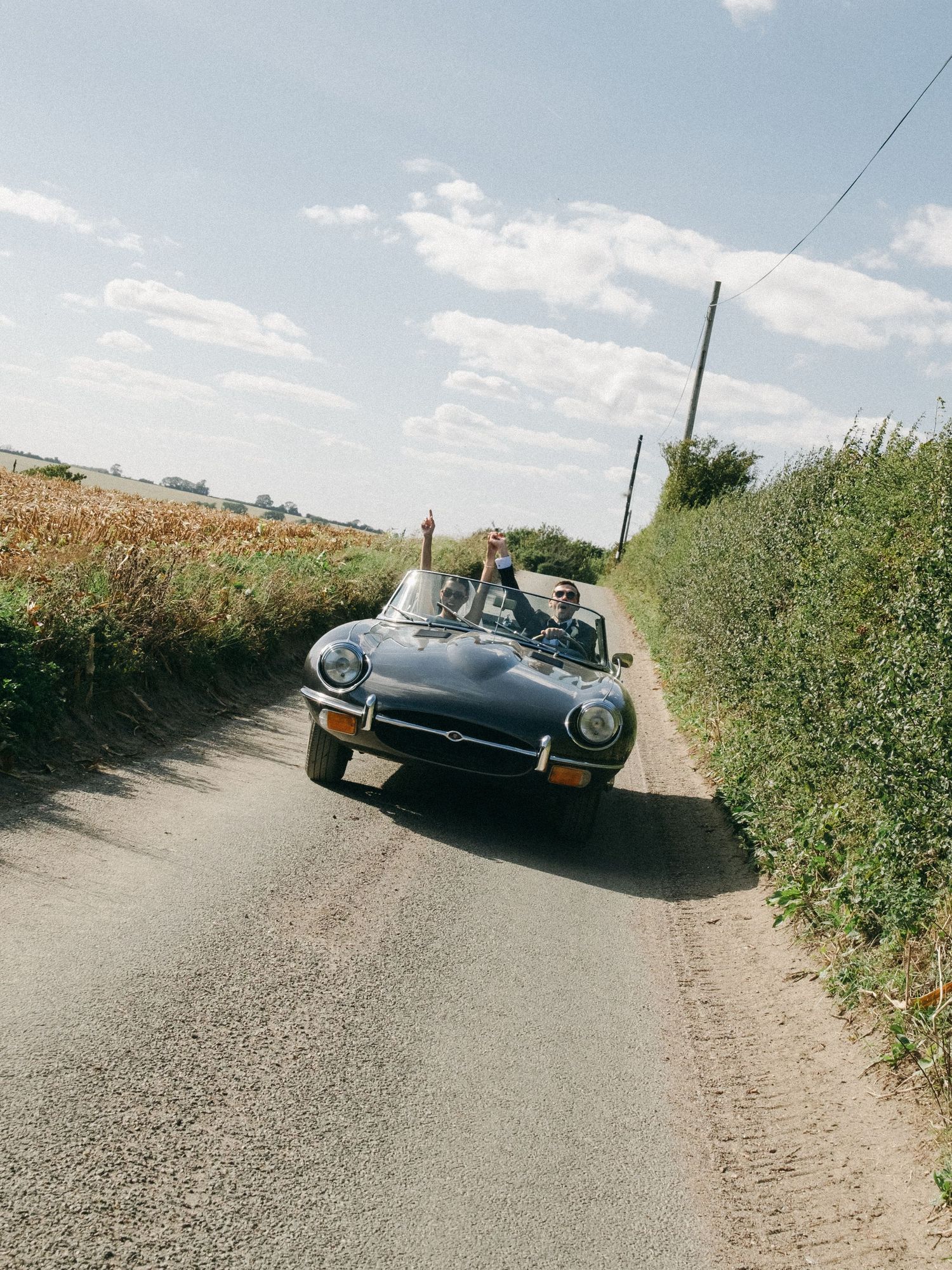 Couple celebrating in a classic black convertible sports car driving down a narrow country lane on a sunny day.