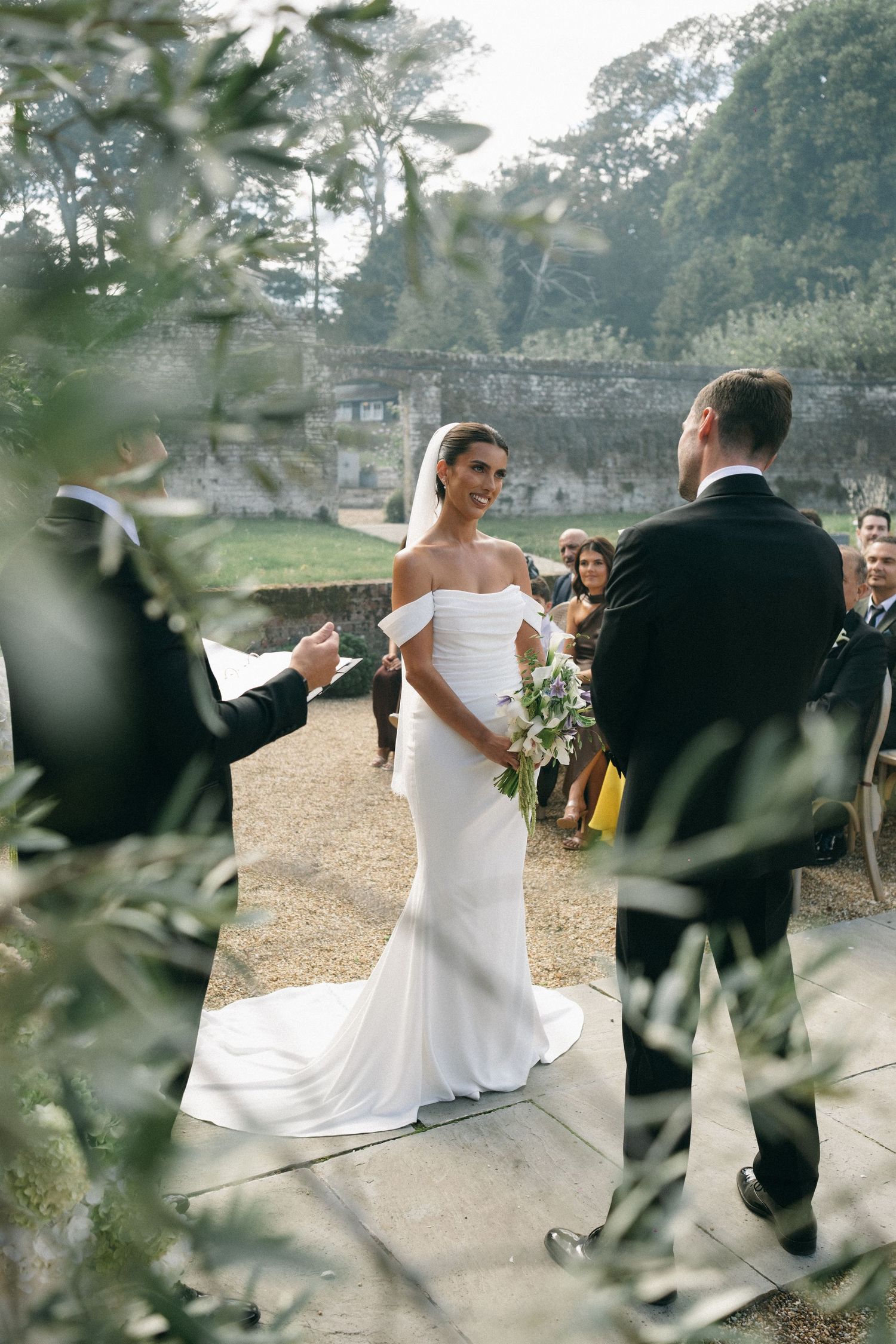 Bride in off-shoulder white gown holds bouquet facing groom at outdoor wedding ceremony with stone walls.
