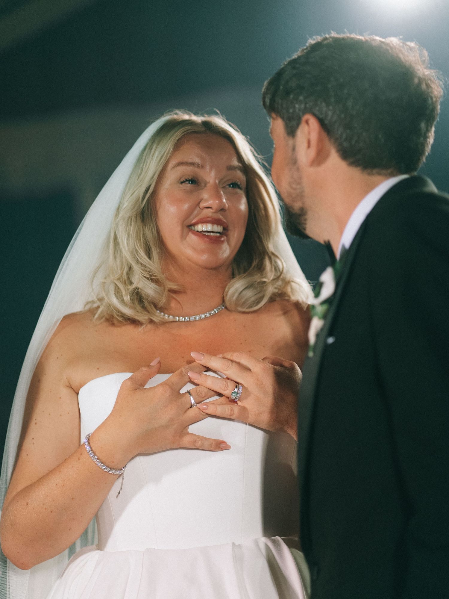 Joyful bride in white strapless gown and veil laughs with groom in dark suit during wedding ceremony.