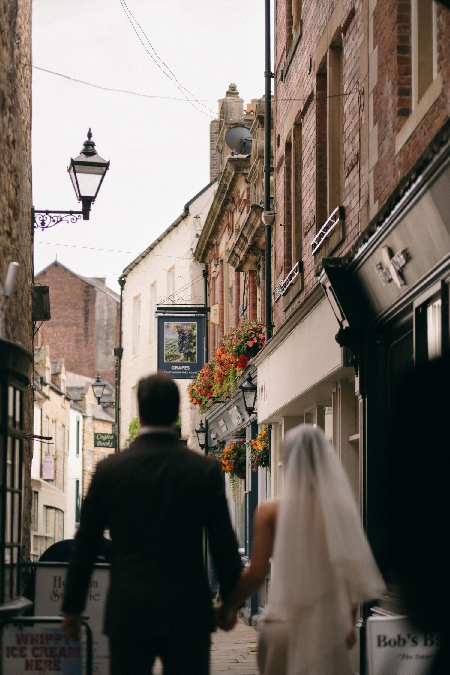 Wedding couple holding hands walking through charming cobblestone street lined with shops and hanging flower baskets.