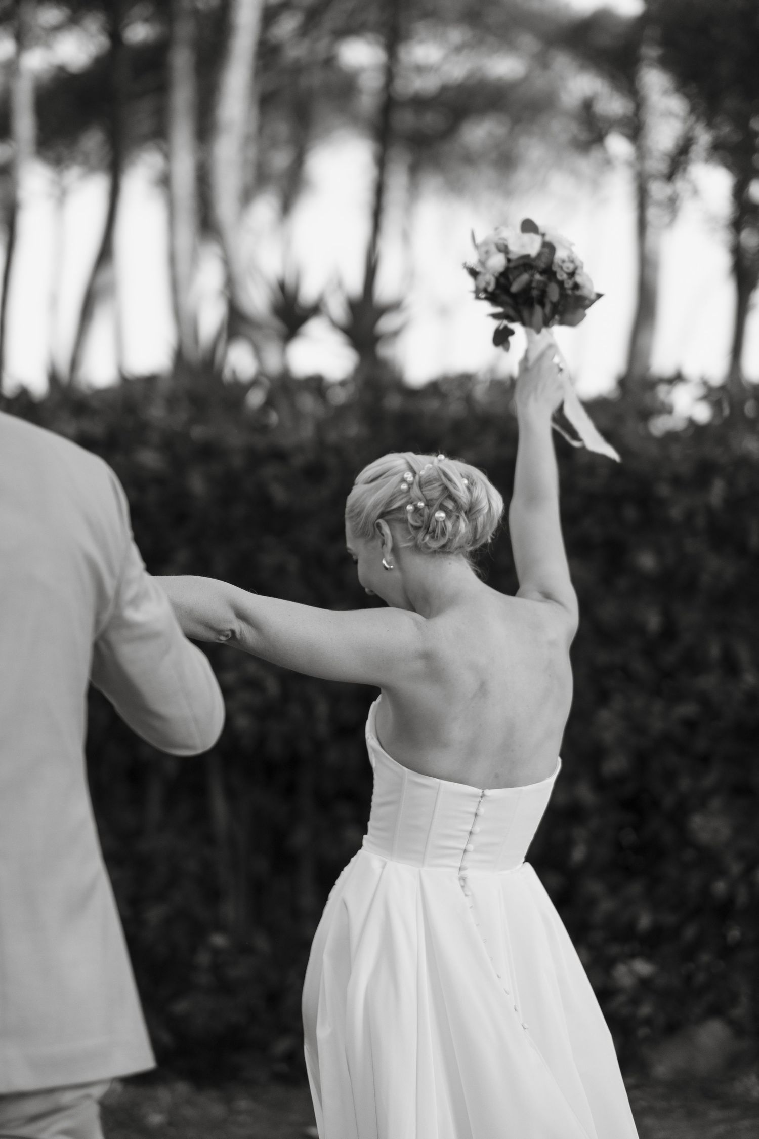 Black and white photo of bride in backless gown raising bouquet while holding grooms hand outdoors.