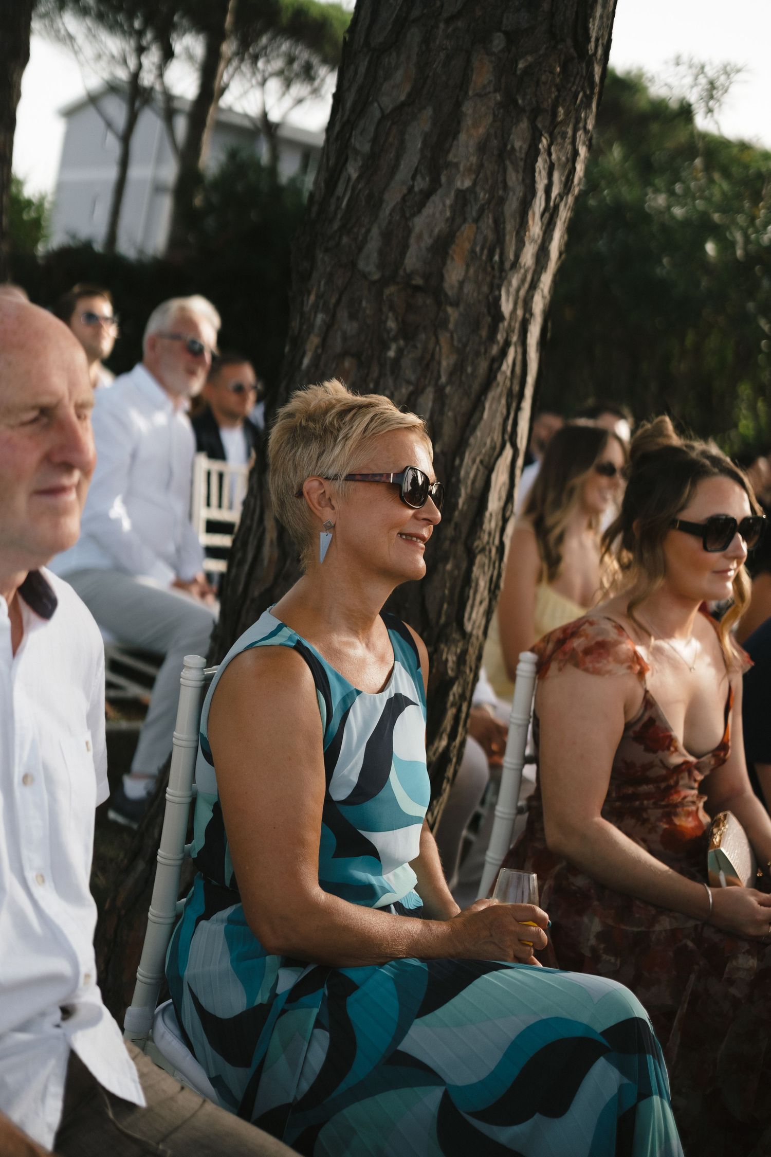 Guests seated outdoors at a wedding ceremony, smiling woman in blue patterned dress wearing sunglasses.