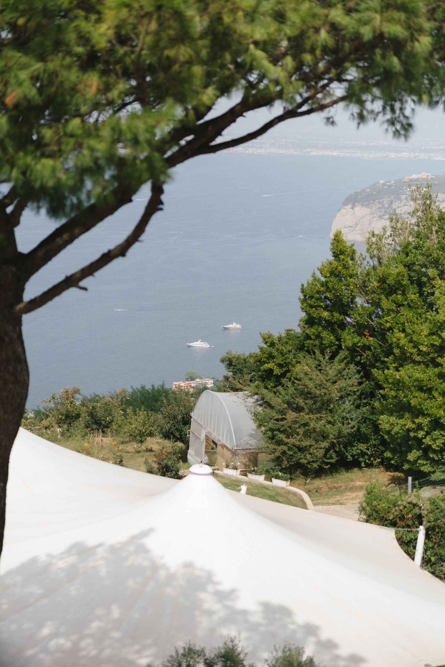 Scenic coastal view from hillside wedding venue with white tent, lush trees, and boats on blue water.