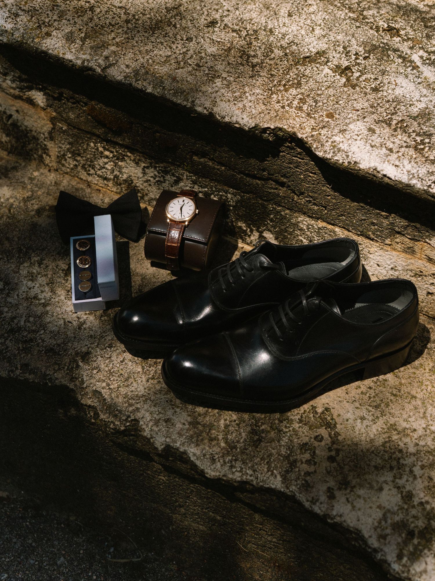 Elegant groom accessories flat lay featuring black Oxford shoes, bow tie, cufflinks, and a brown leather watch box.
