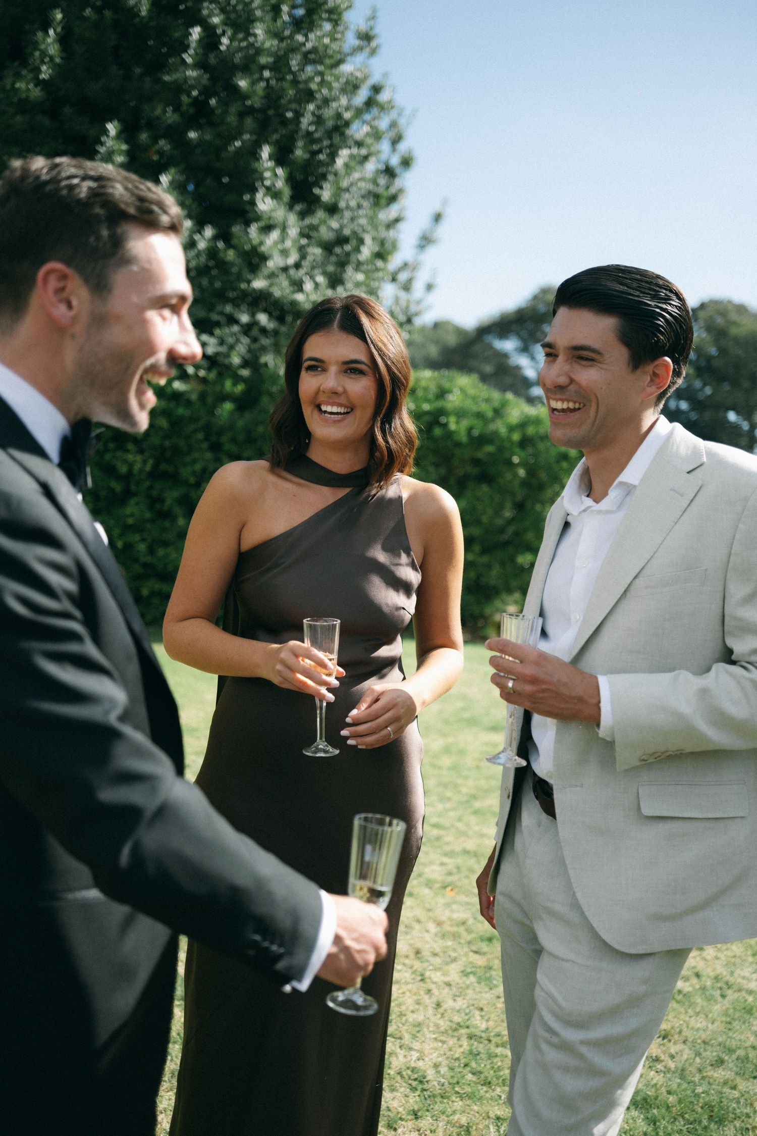 Three elegantly dressed guests socializing outdoors at a wedding, holding champagne glasses on a sunny day.