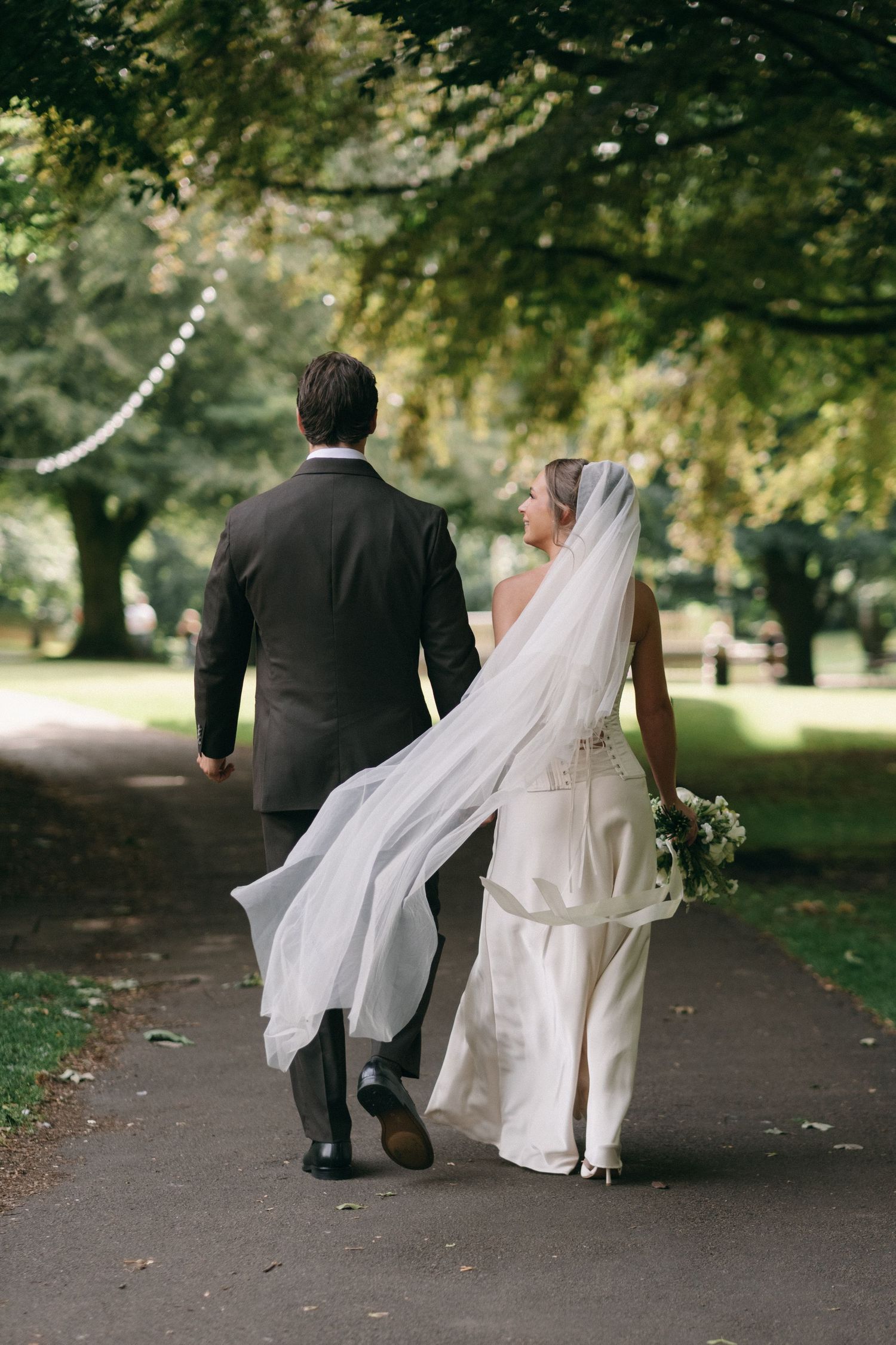 Bride and groom walk hand in hand down tree-lined path, bride's veil flowing in the breeze.