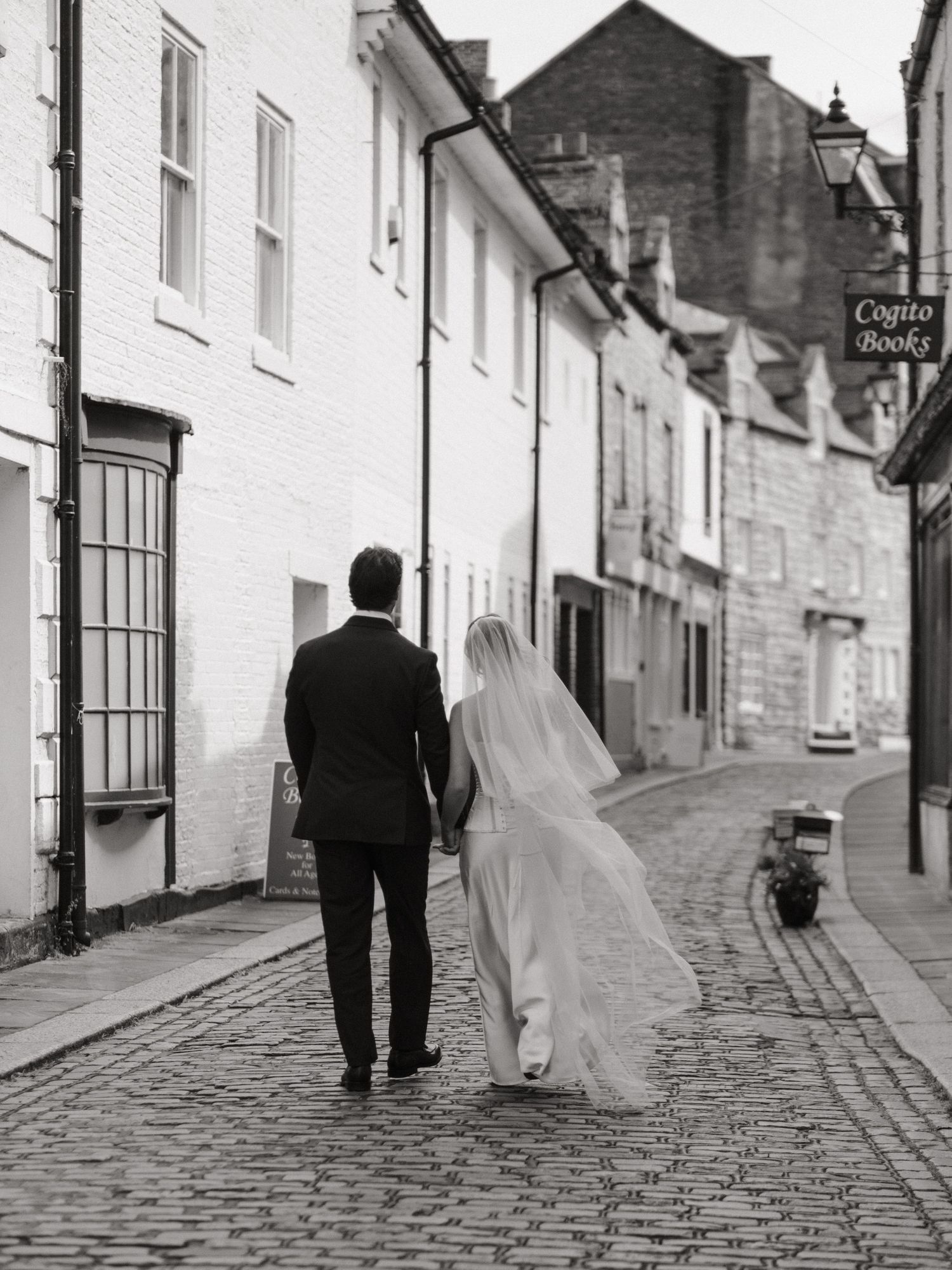 Black and white photo of bride and groom walking hand in hand down historic cobblestone street with veil flowing.
