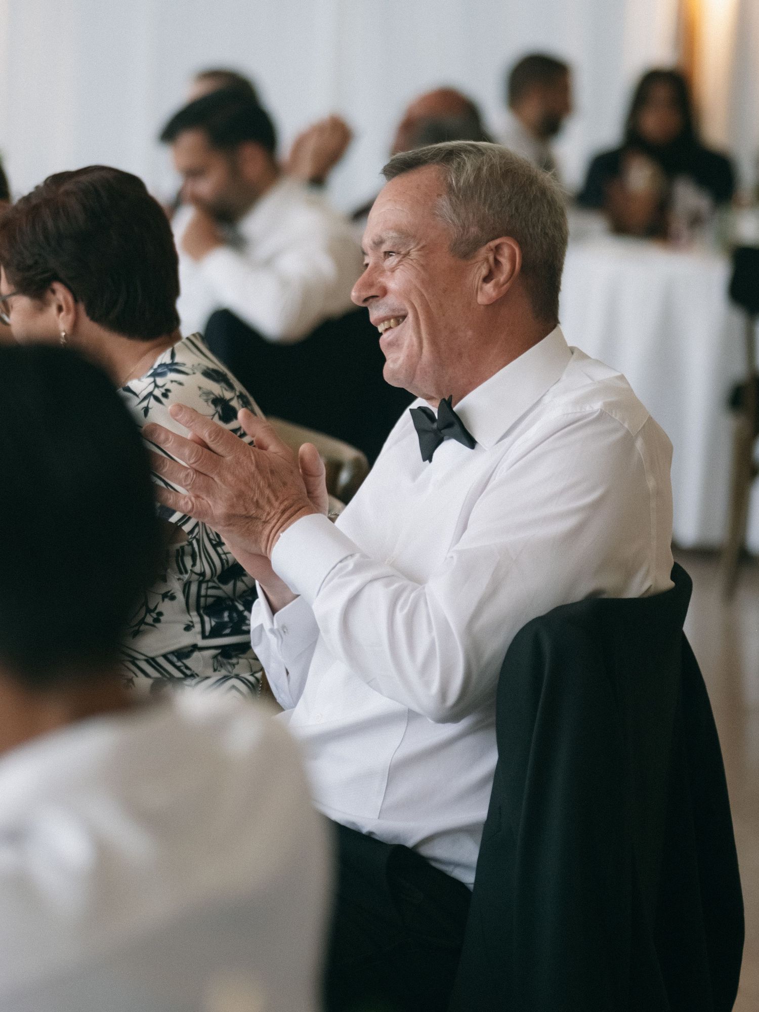 Elderly man in bow tie clapping and smiling at a wedding reception.