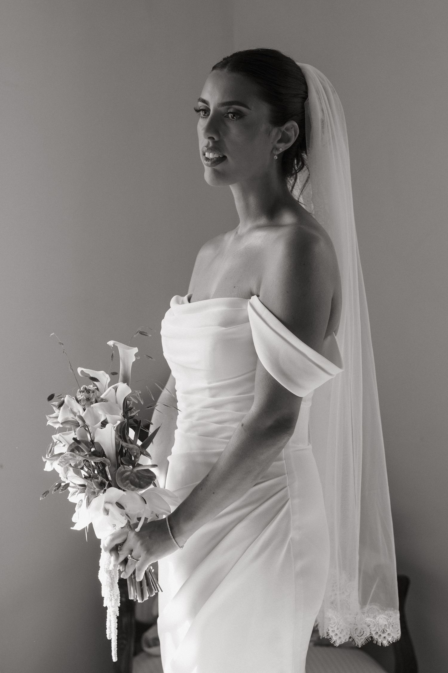 Black and white portrait of a bride in an off-shoulder gown holding a floral bouquet with a veil.