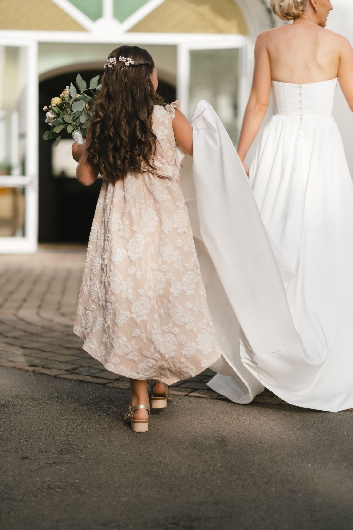 Flower girl in lace dress holding bouquet walking beside bride in white gown.