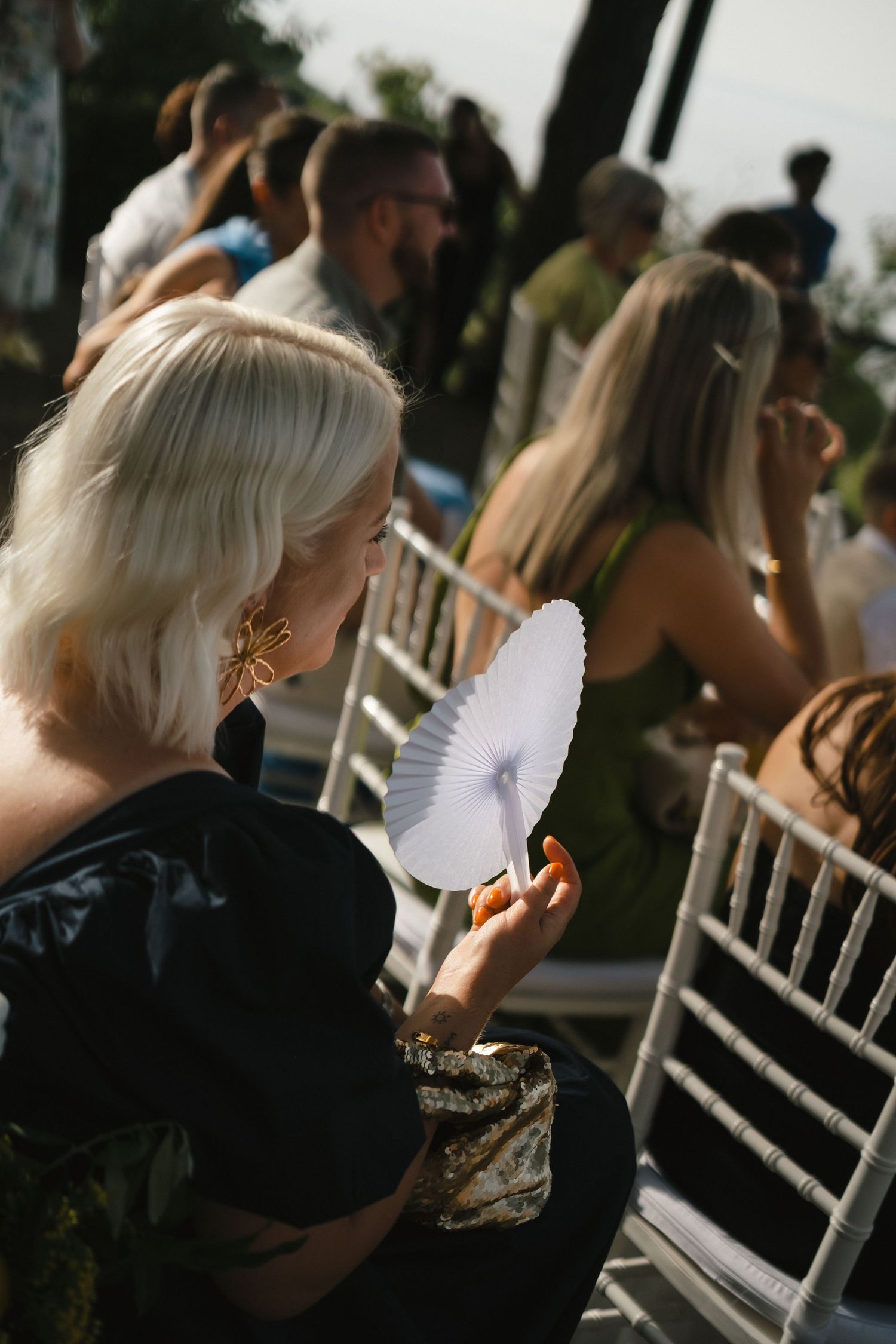 Blonde woman in black dress holding white paper fan at outdoor wedding ceremony.