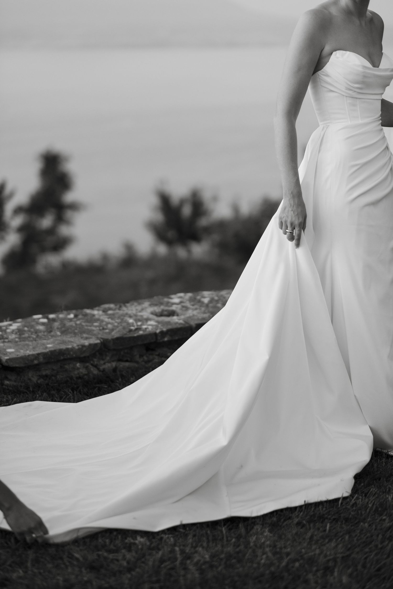Black and white photo of bride in strapless gown with flowing train outdoors.
