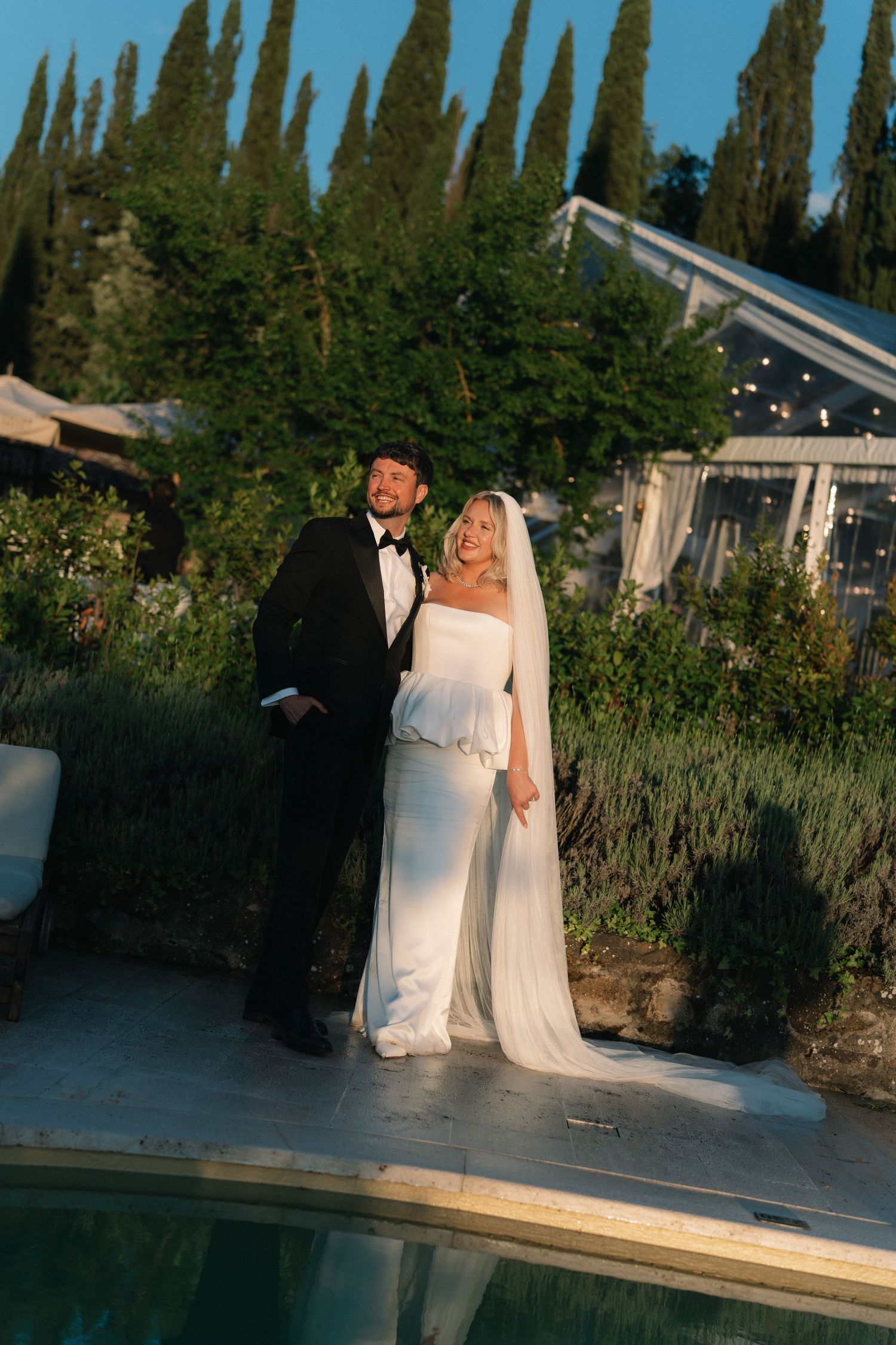 Bride in white gown and veil stands with groom in black tuxedo by a pool surrounded by cypress trees at golden hour.