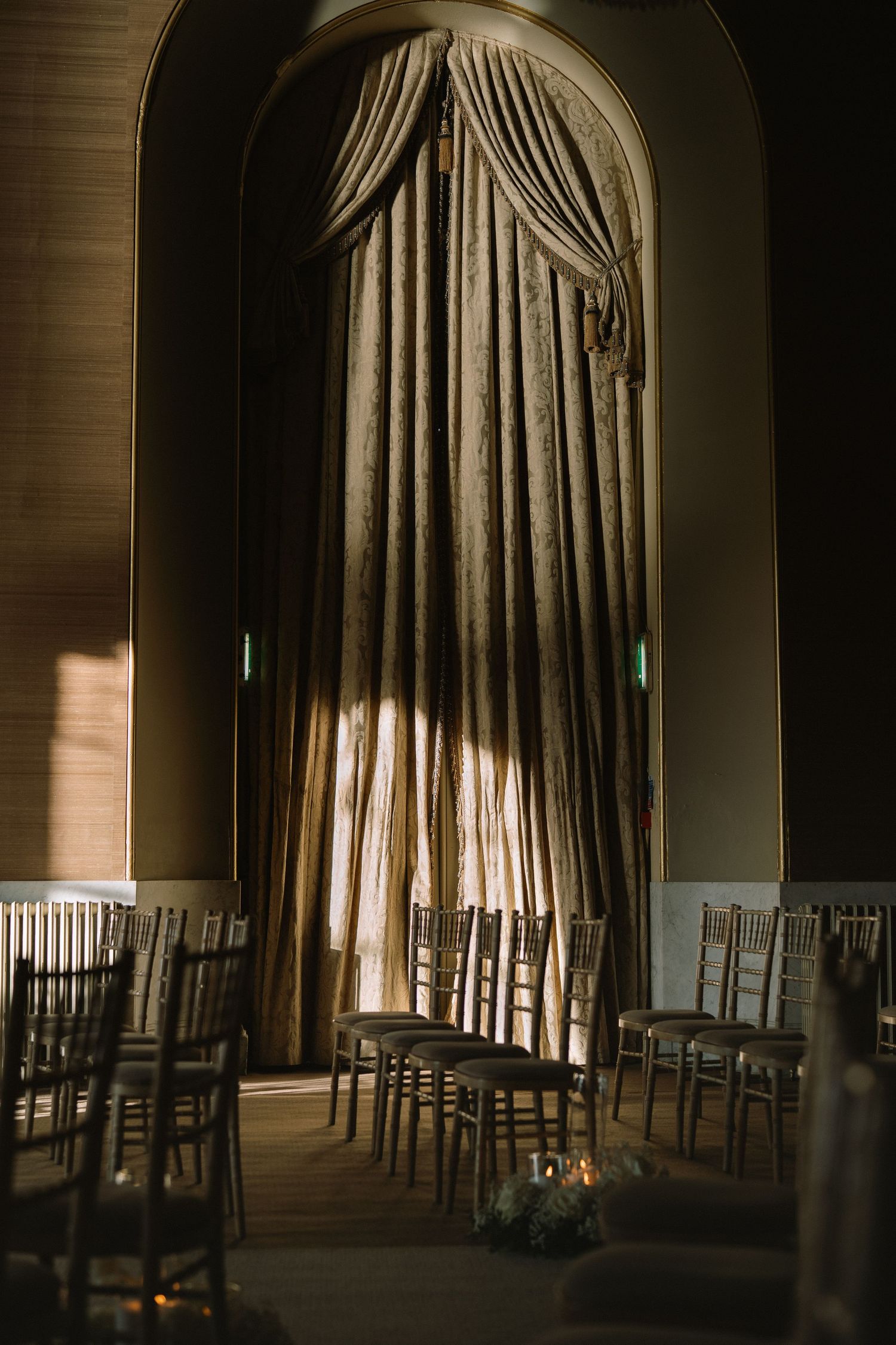 Elegant wedding venue with chiavari chairs and dramatic floor-length curtains in an arched window setting.