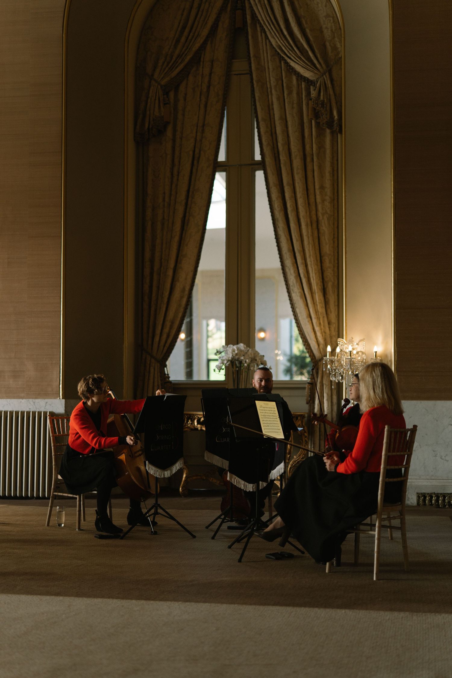 String quartet in red attire performing in an elegant ballroom with tall draped windows and candlelight.