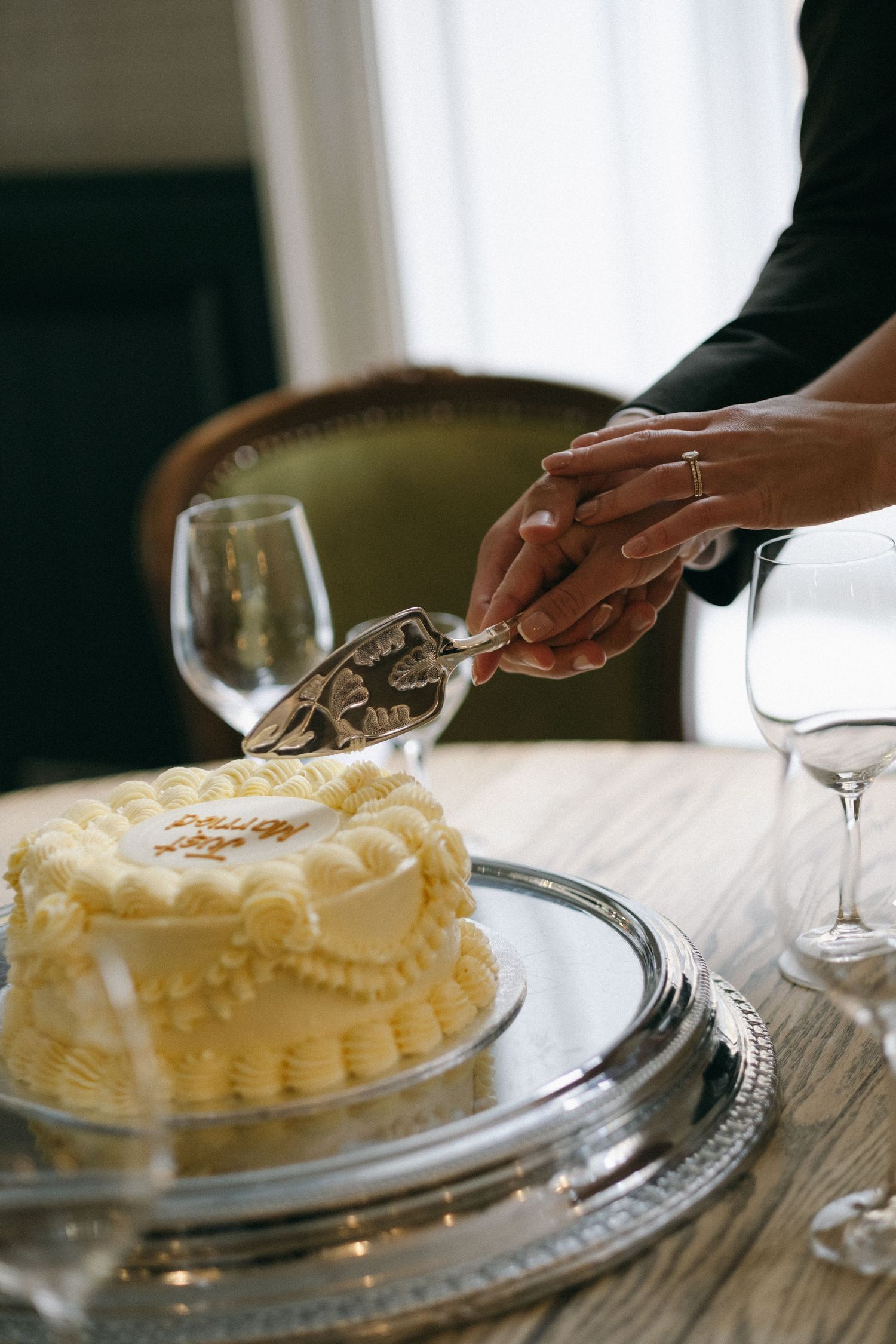 Couple cutting elegant white frosted wedding cake on silver platter with ornate cake server.