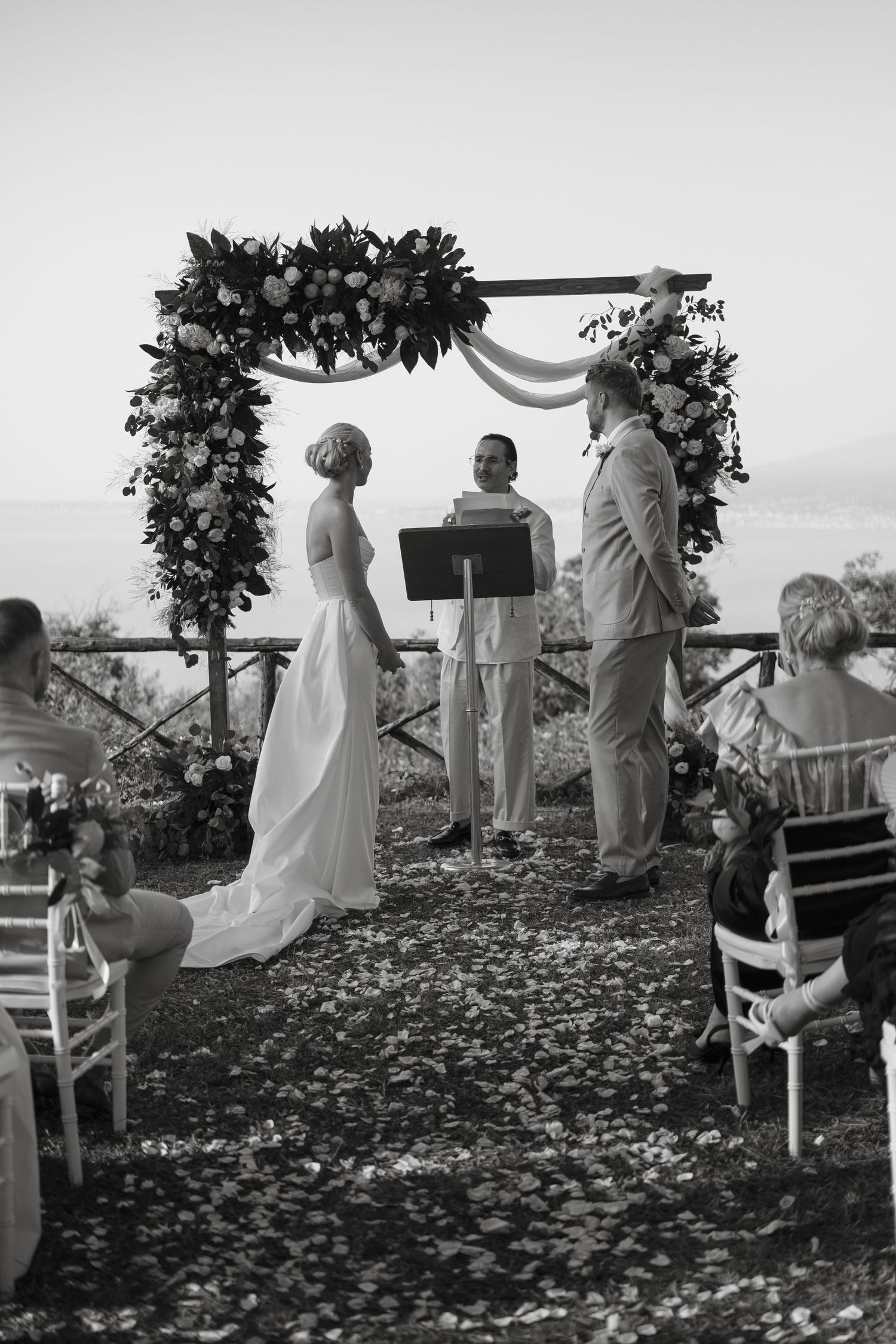 Black and white photo of bride and groom exchanging vows under a floral arch at an outdoor wedding ceremony.