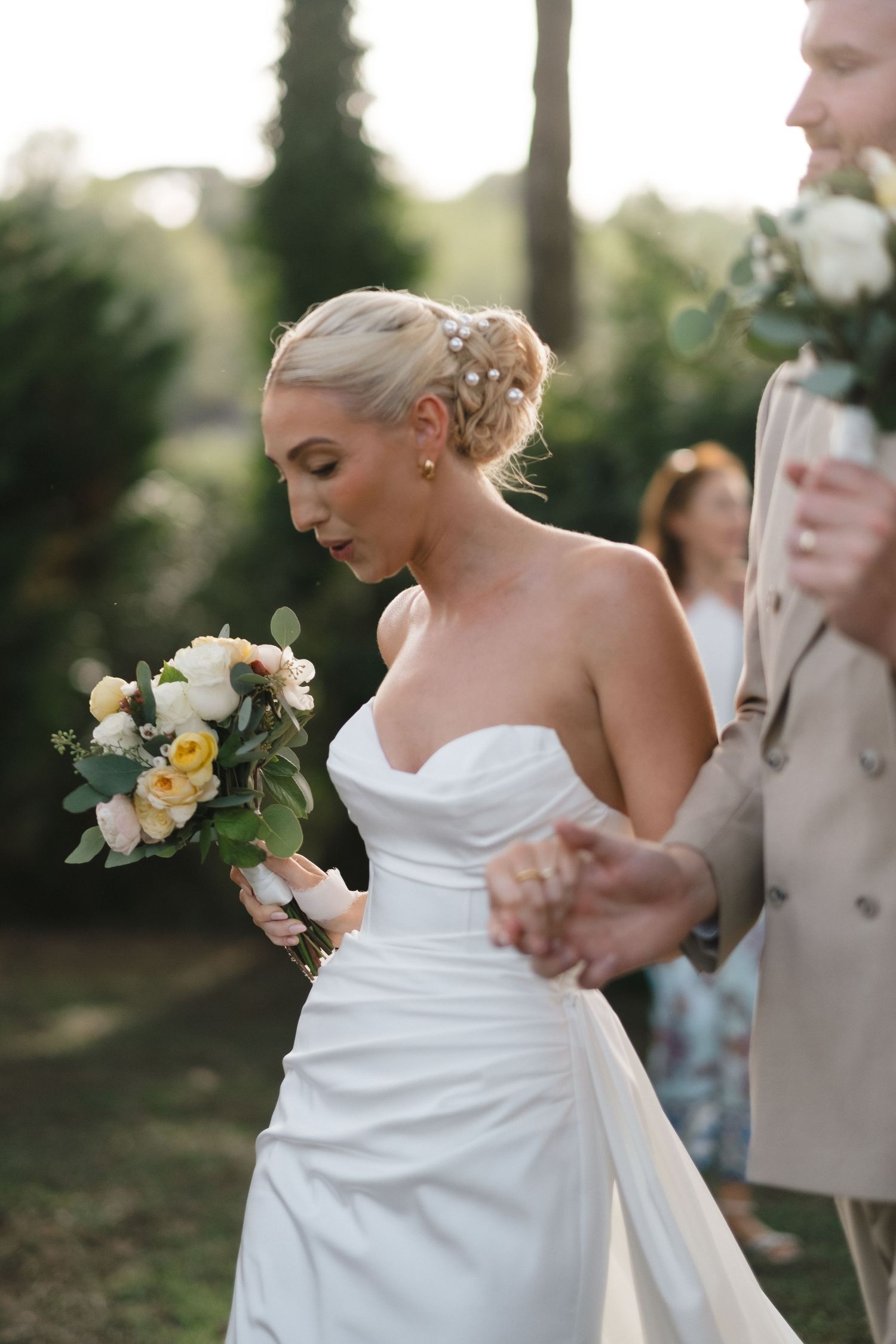 Blonde bride in strapless white gown holds yellow and white bouquet while holding grooms hand outdoors.