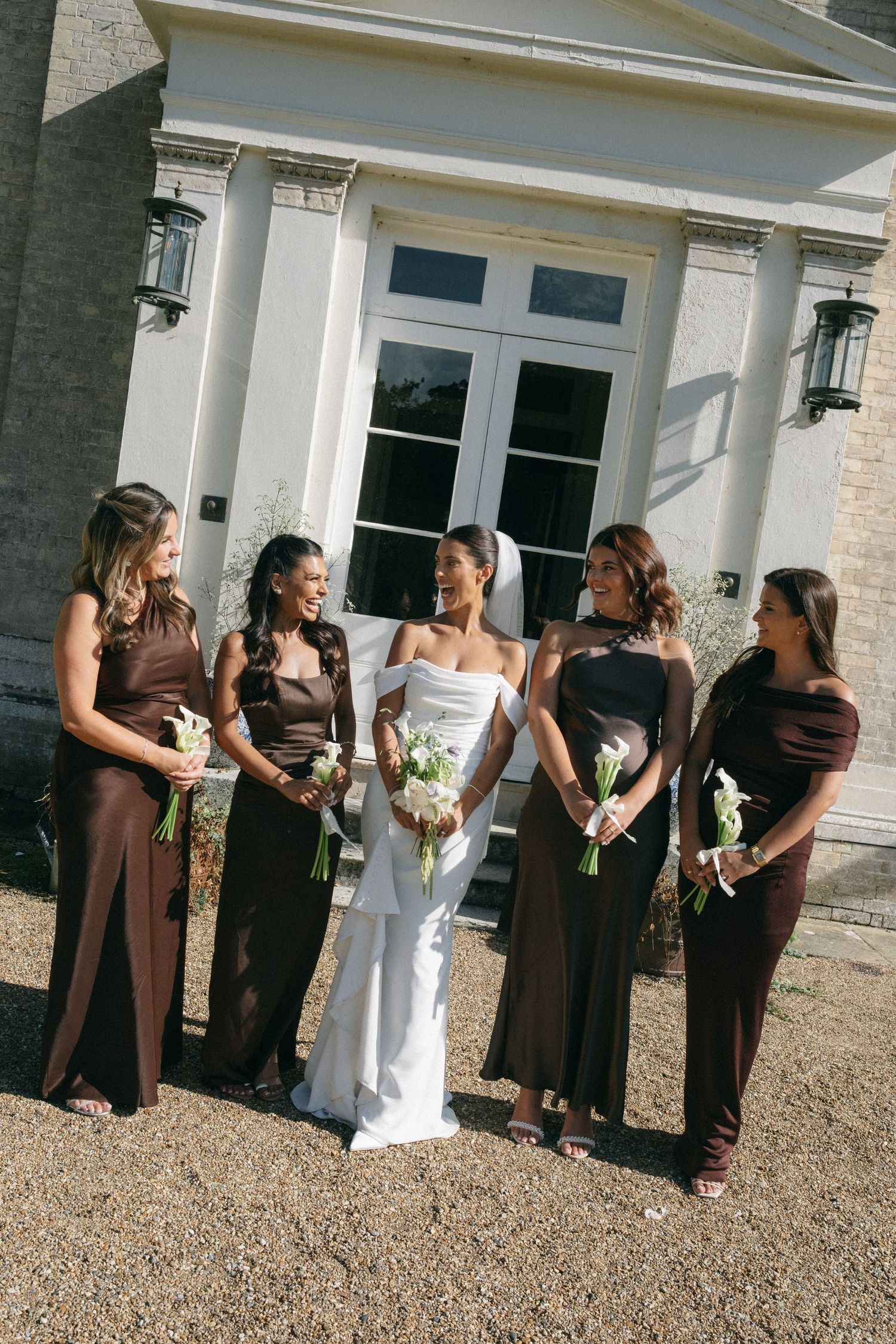 Bride in white gown with four bridesmaids in brown dresses holding white bouquets outside elegant venue.