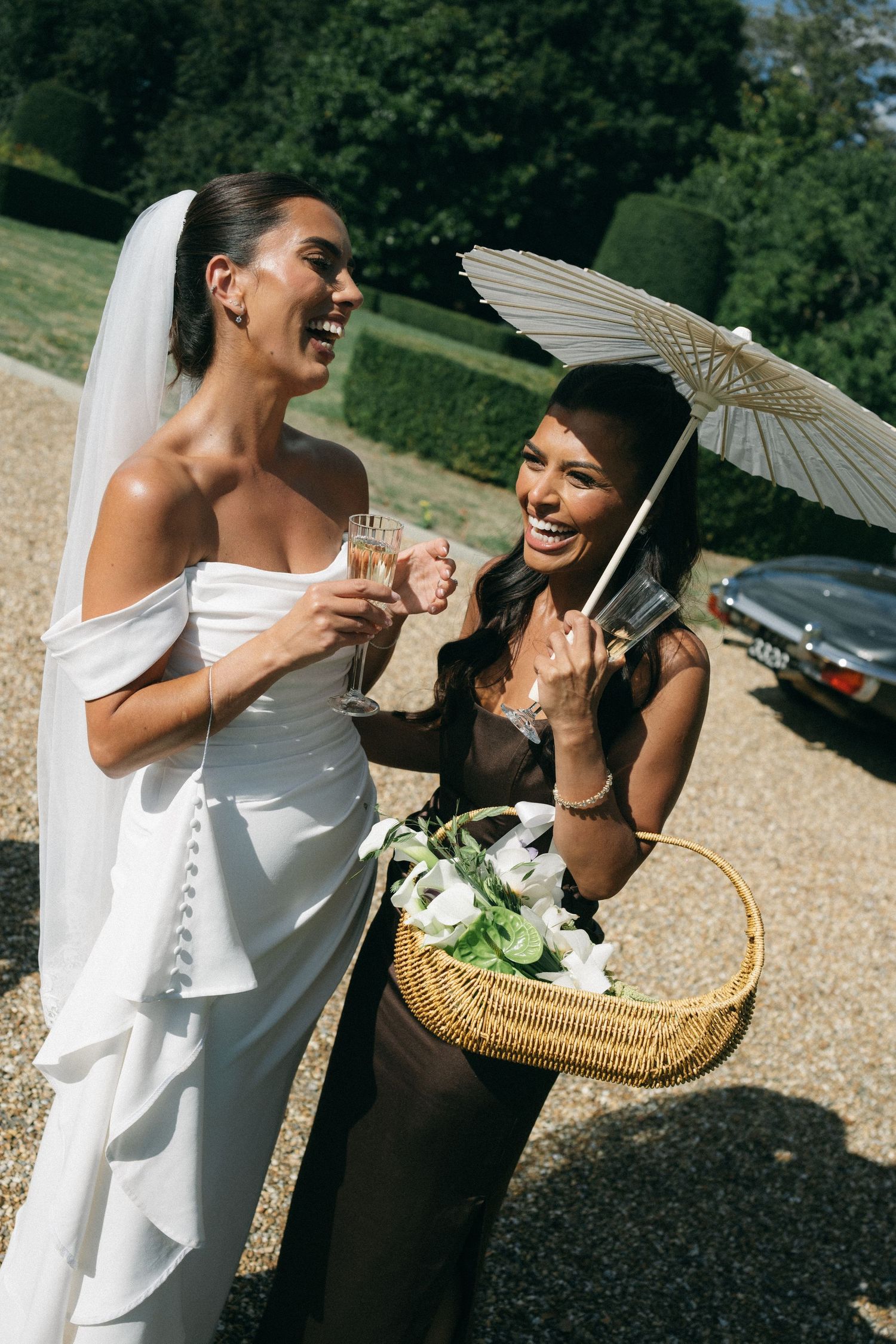Bride in white gown and veil laughs with bridesmaid holding parasol and flower basket at outdoor wedding.