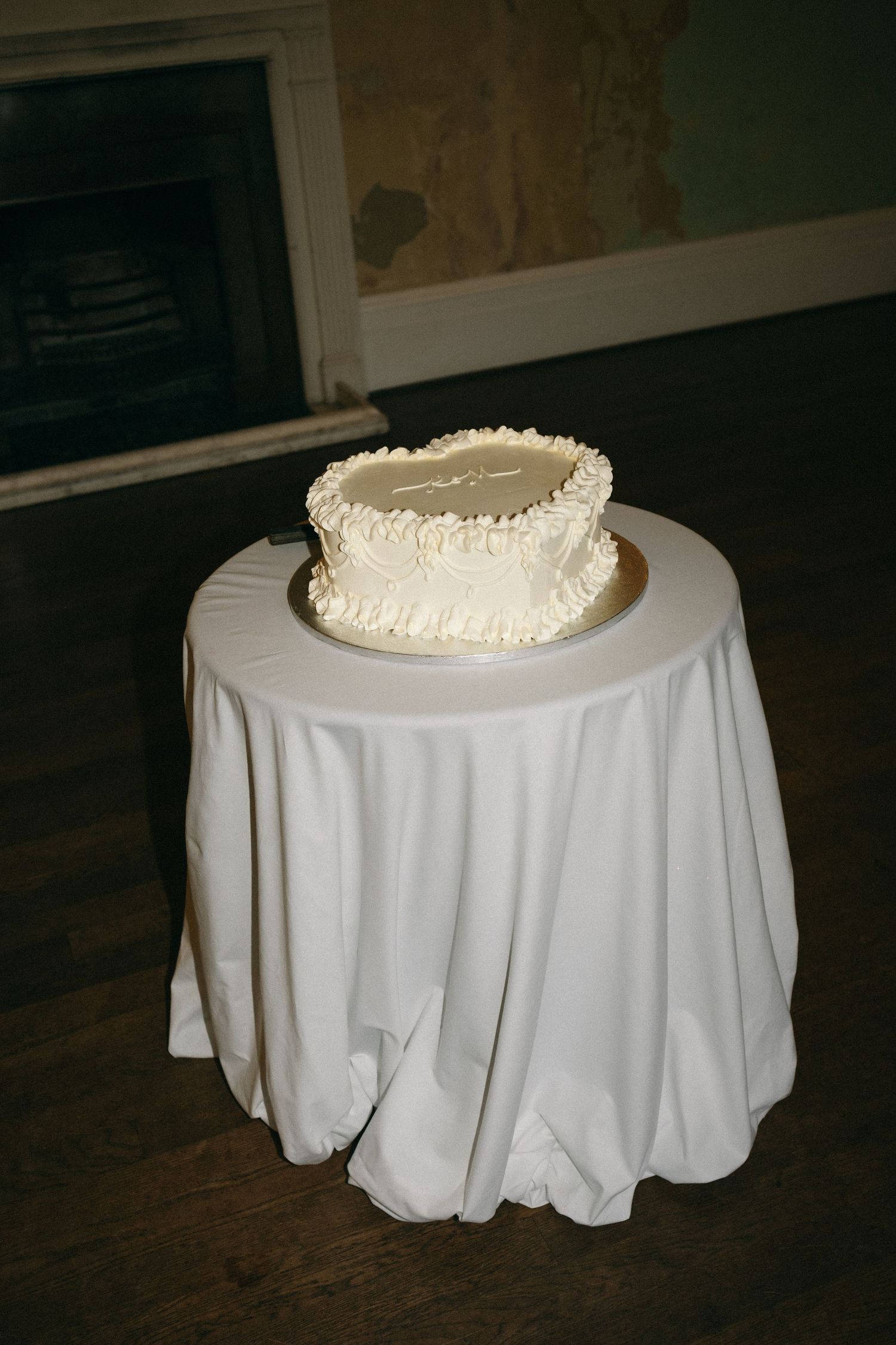 Single-tier white wedding cake with ruffled frosting on a white draped table near a fireplace.