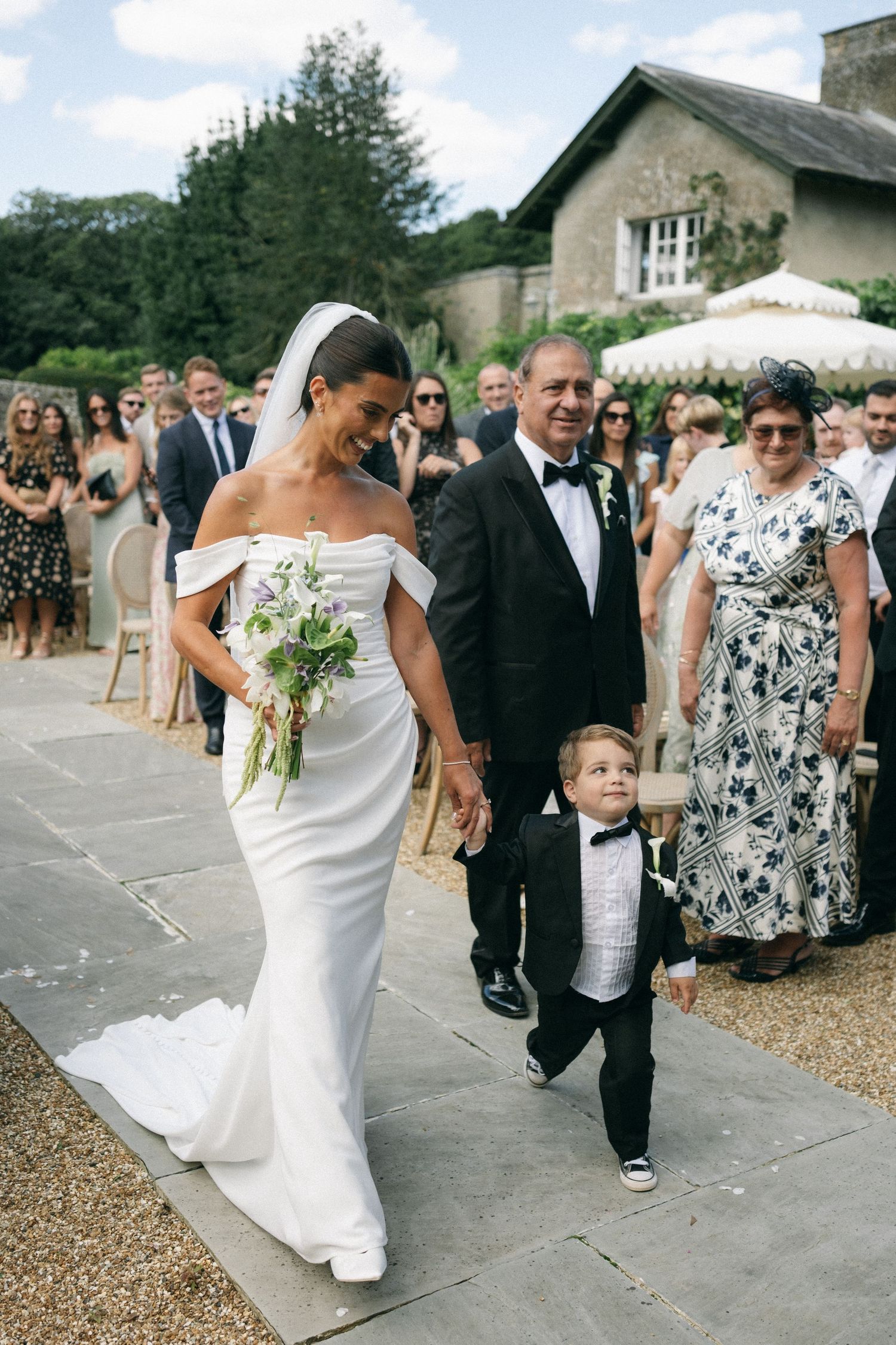 Bride in white off-shoulder gown walks down the aisle with a man in tuxedo and young ring bearer at outdoor wedding.
