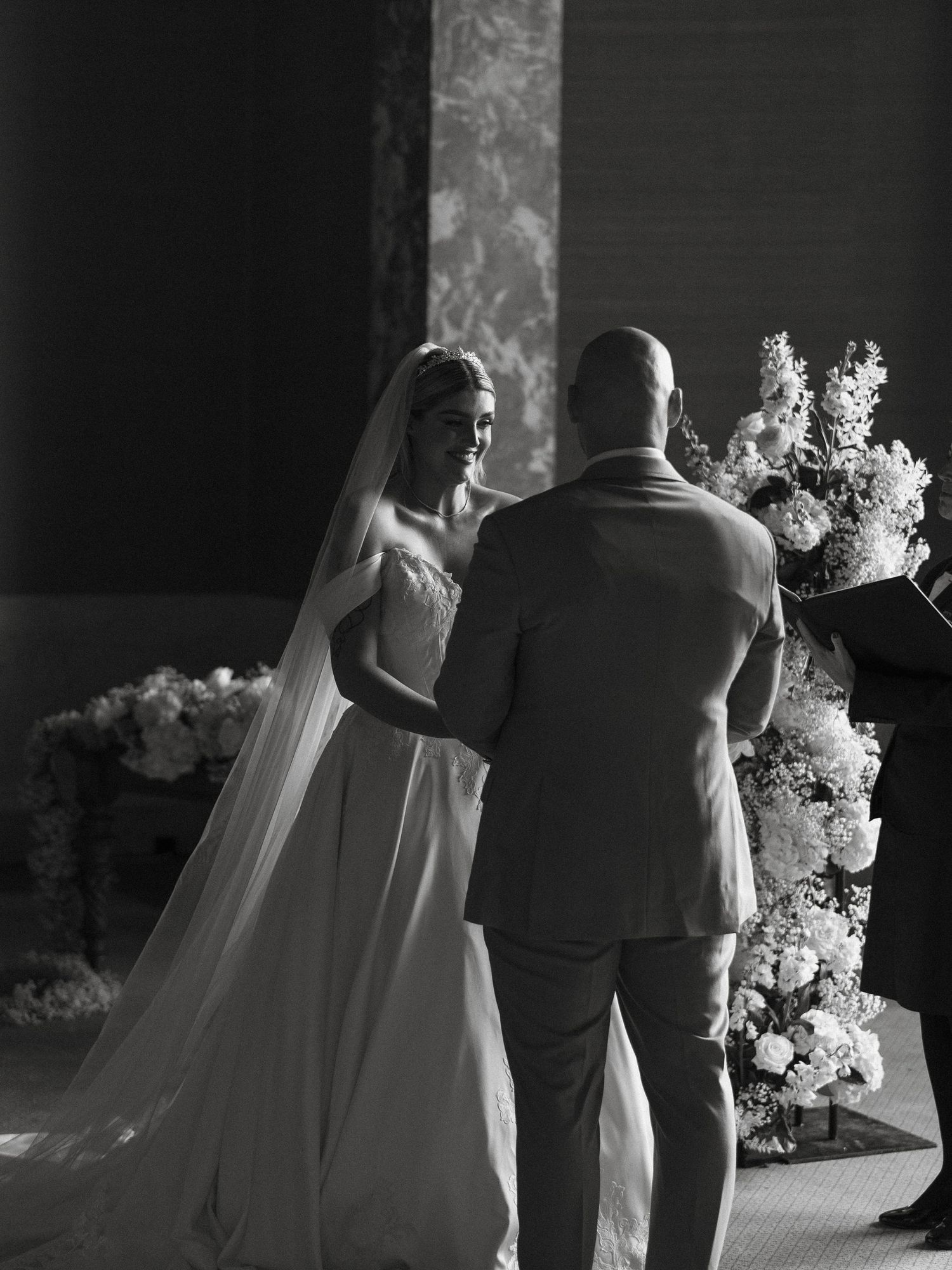 Black and white photo of a bride and groom exchanging vows at a floral-decorated wedding altar.