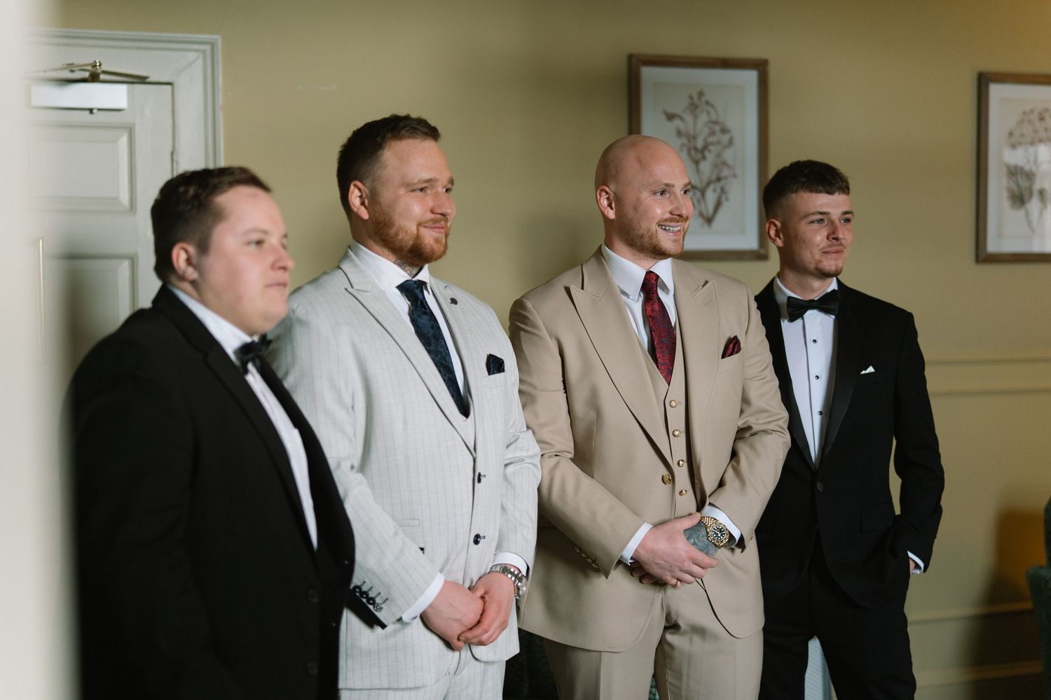 Four groomsmen in formal suits pose together indoors at a wedding ceremony venue.