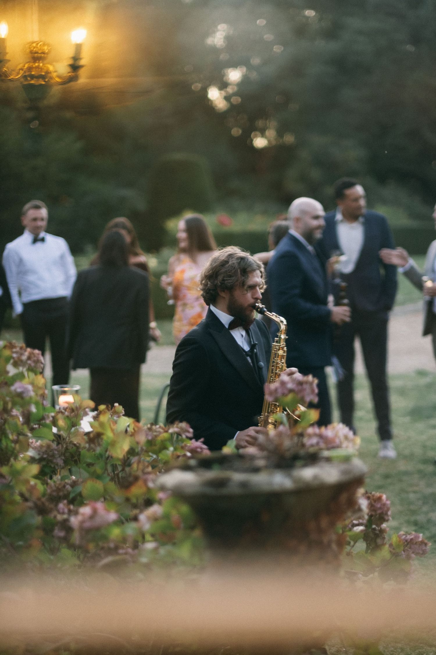 Saxophonist in black tuxedo performs outdoors at elegant garden wedding reception at dusk.
