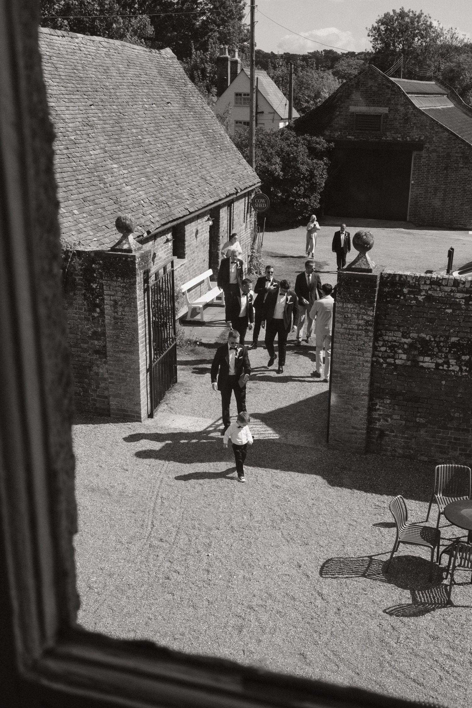 Black and white aerial view of wedding guests gathering in a rustic courtyard surrounded by brick buildings and lush trees.
