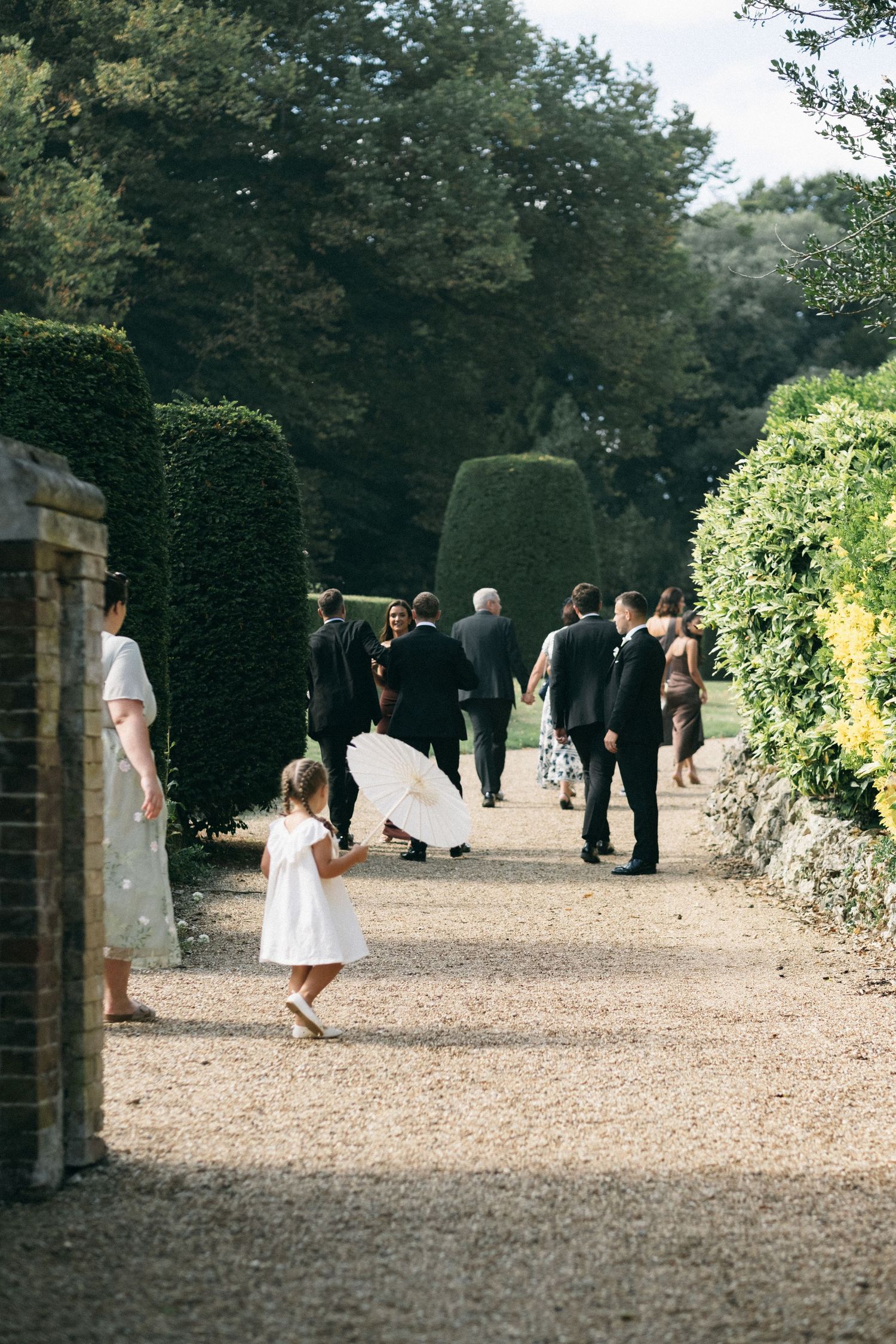 A young girl in a white dress carries a parasol along a garden path at a wedding celebration.