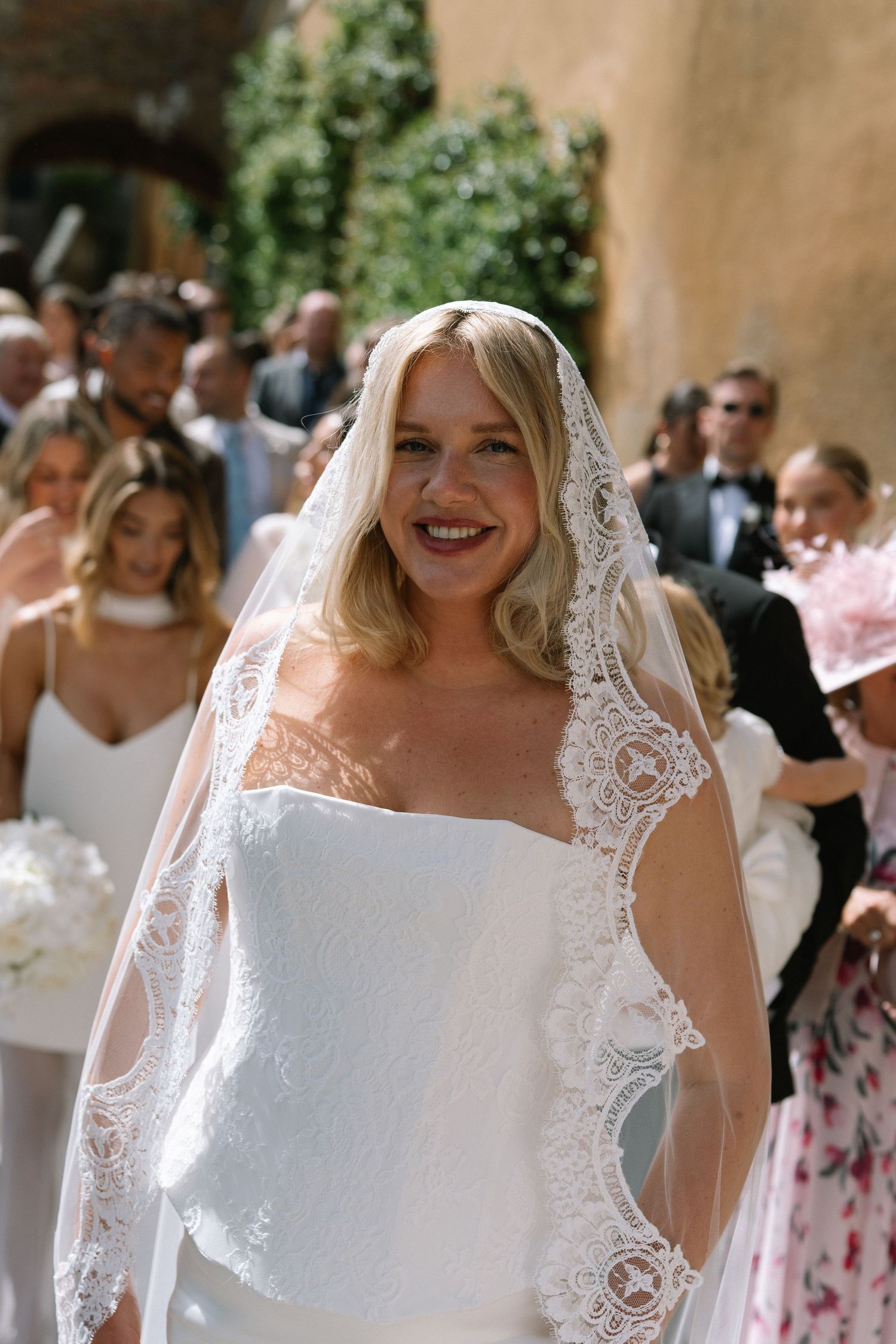 Smiling blonde bride in strapless gown with ornate lace-edged veil walks among wedding guests outdoors.