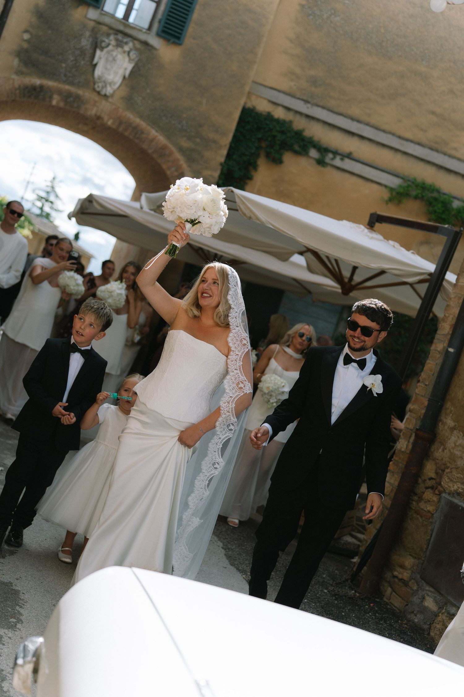 Joyful bride raising her white bouquet while walking with groom in sunglasses through a Tuscan villa archway.