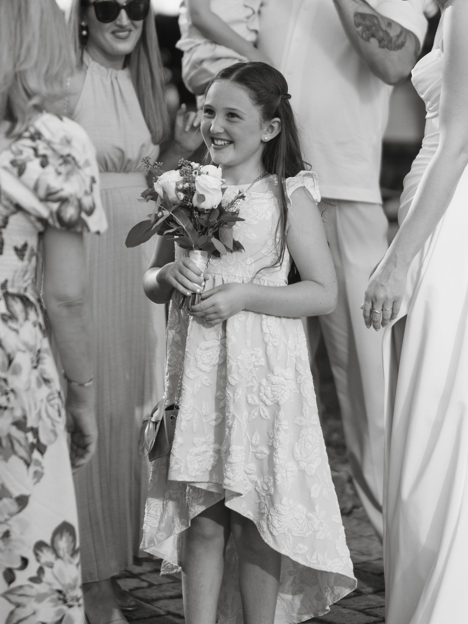 Smiling young flower girl in white lace dress holds bouquet of roses at outdoor wedding ceremony.