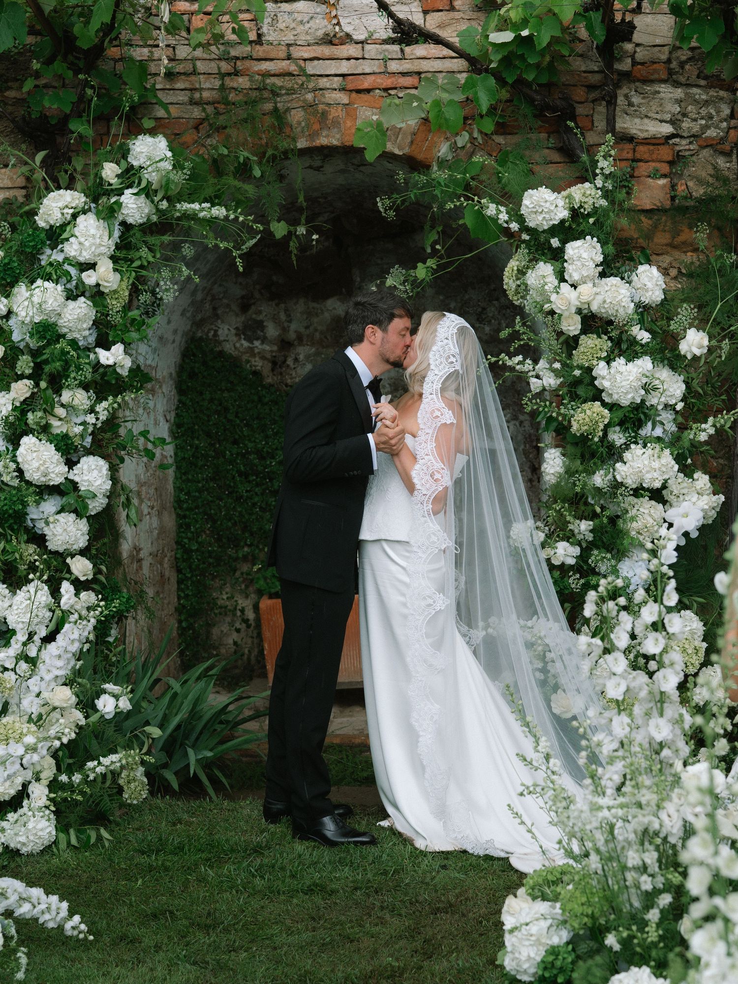 Newlyweds share their first kiss under a stunning white floral arch at an outdoor wedding.