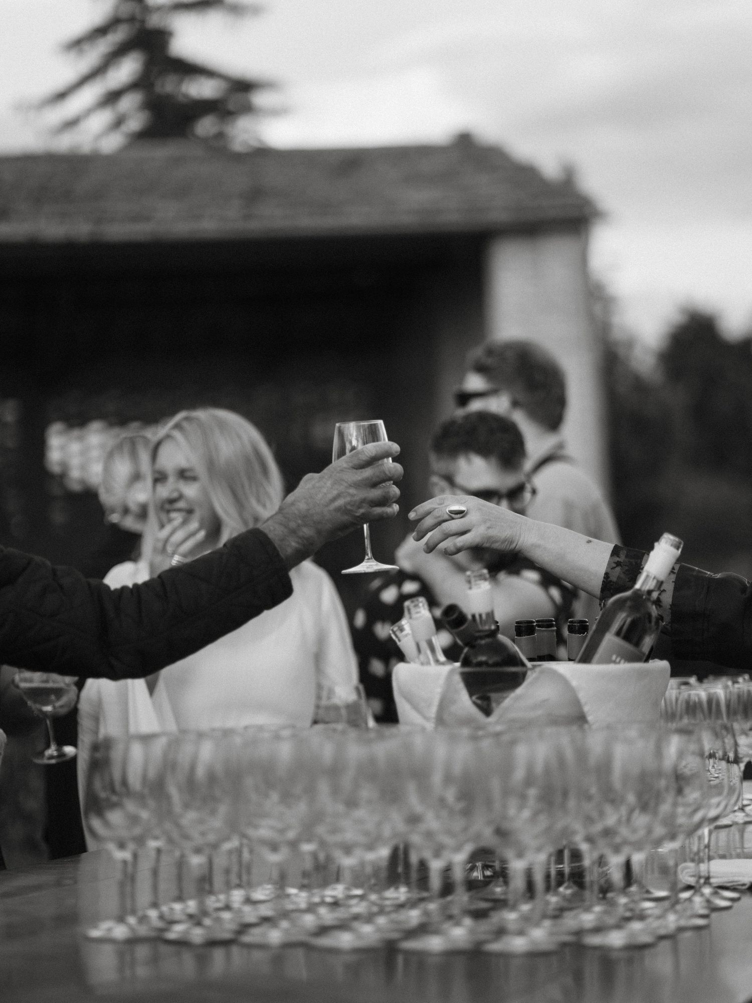 Black and white photo of guests raising champagne glasses in a celebratory toast at an outdoor wedding.