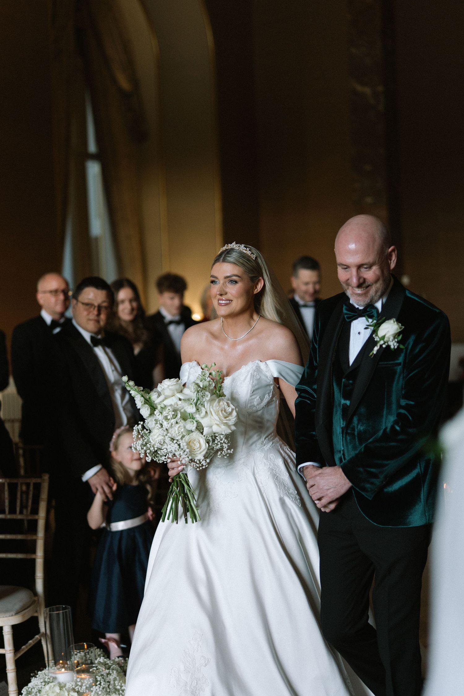 Bride in off-shoulder gown holding white bouquet walks down aisle with man in green velvet tuxedo.