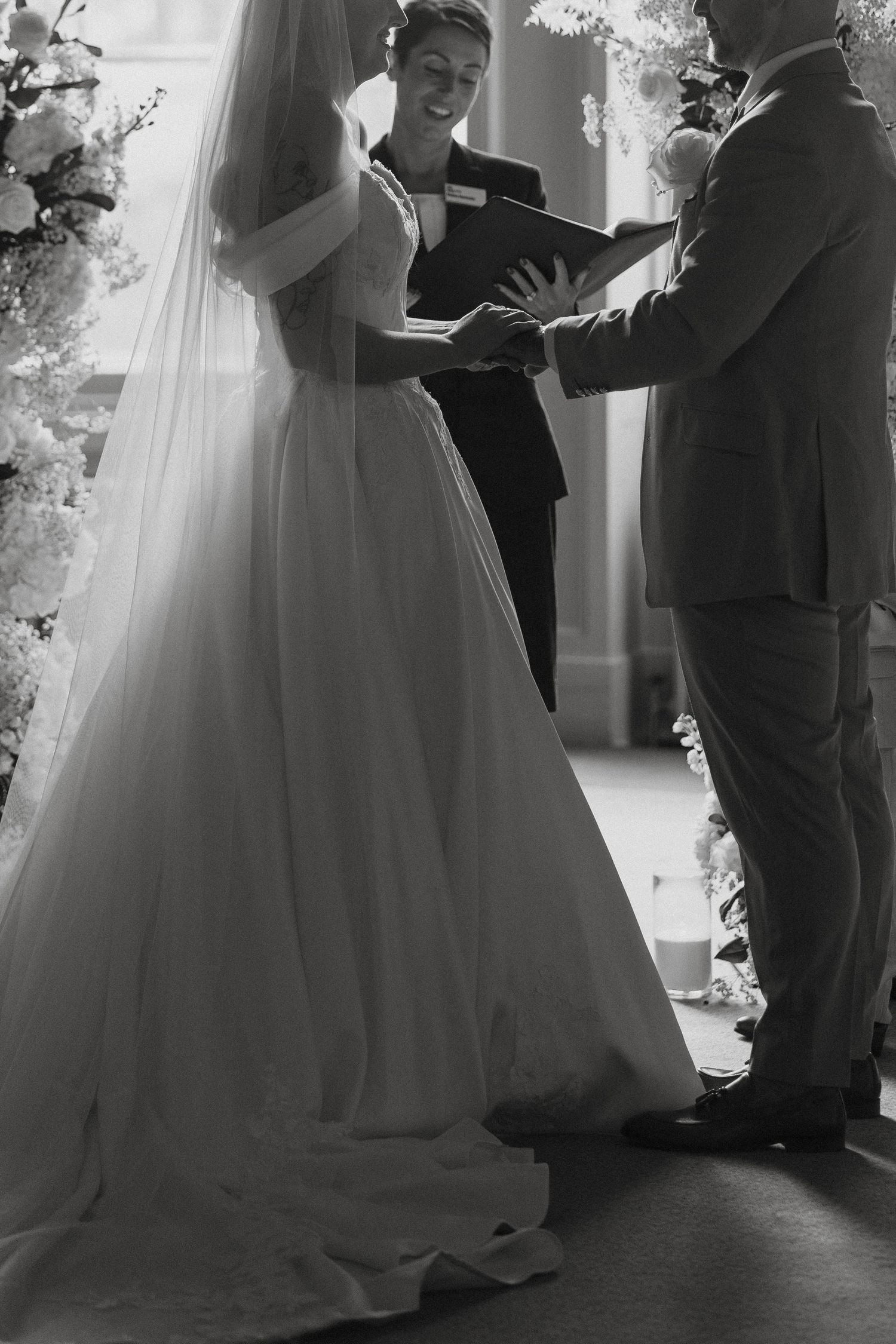 Black and white wedding ceremony photo of bride in flowing gown and veil exchanging vows with officiant.