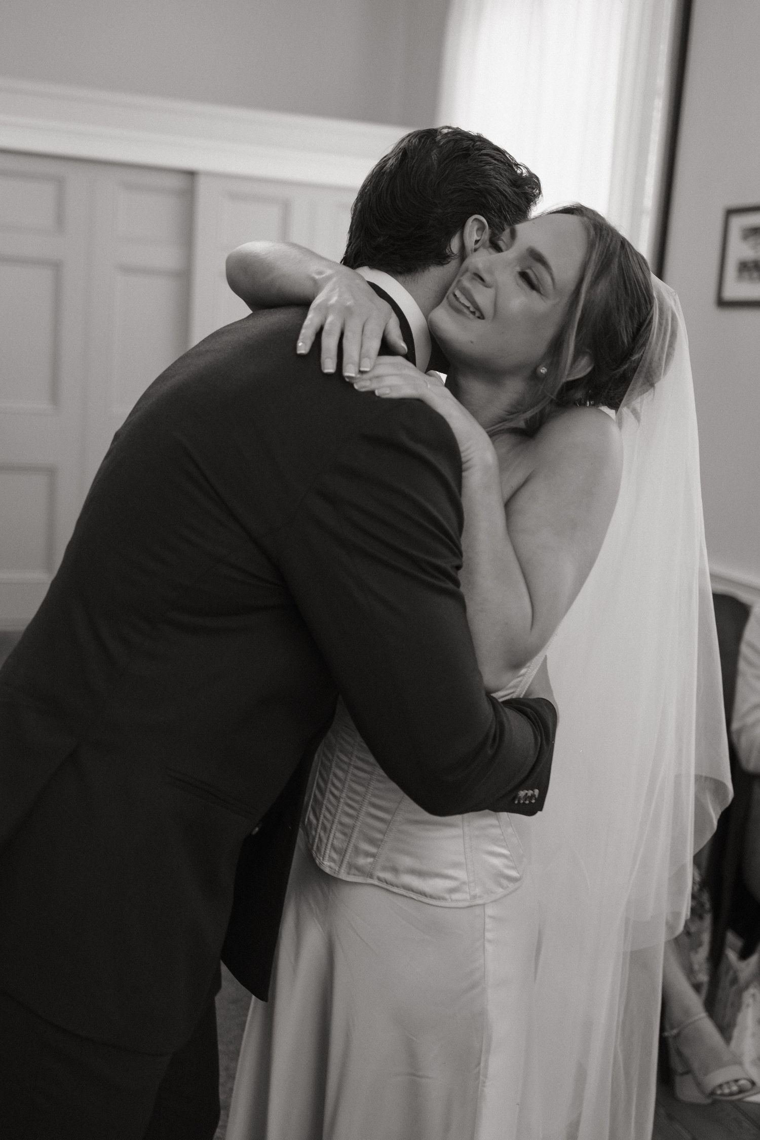 Black and white photo of a bride in a veil embracing a groom in a suit, smiling joyfully during their wedding day.