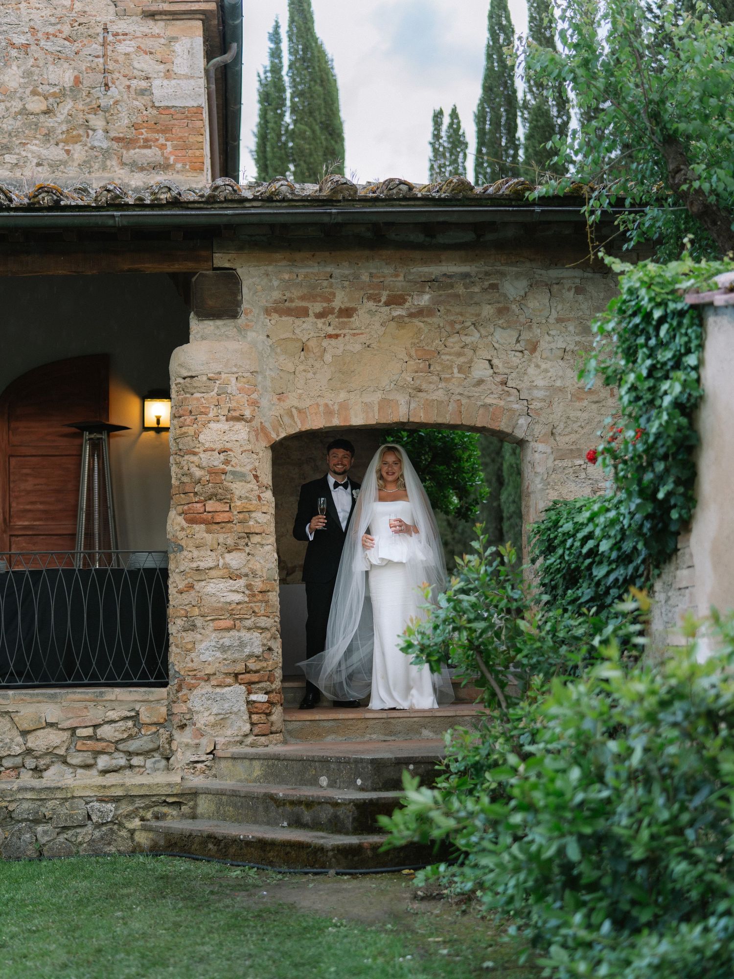Newlywed couple in wedding attire emerging through a stone archway at a rustic Tuscan villa wedding venue.