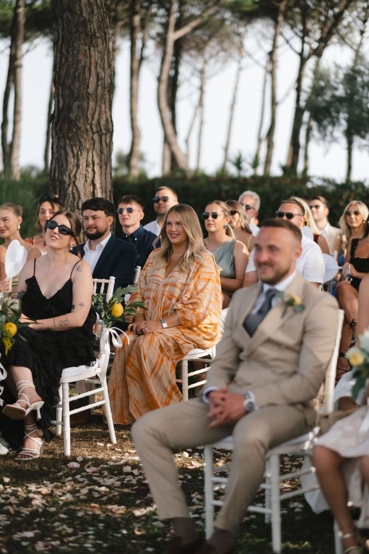Wedding guests seated outdoors under pine trees, watching a ceremony, with a man in a beige suit in the foreground.