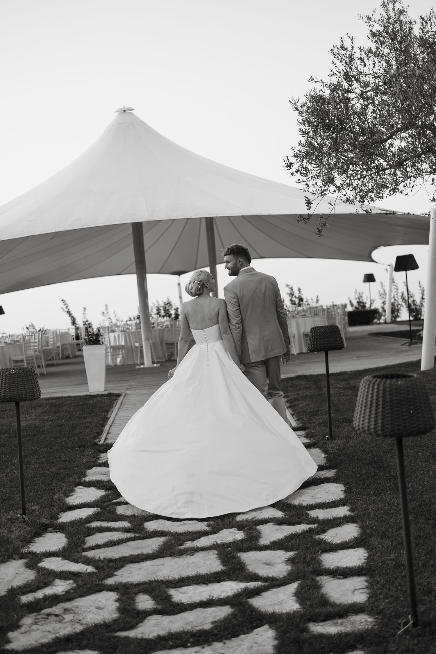 Black and white photo of bride and groom walking toward elegant outdoor wedding tent on stone path.