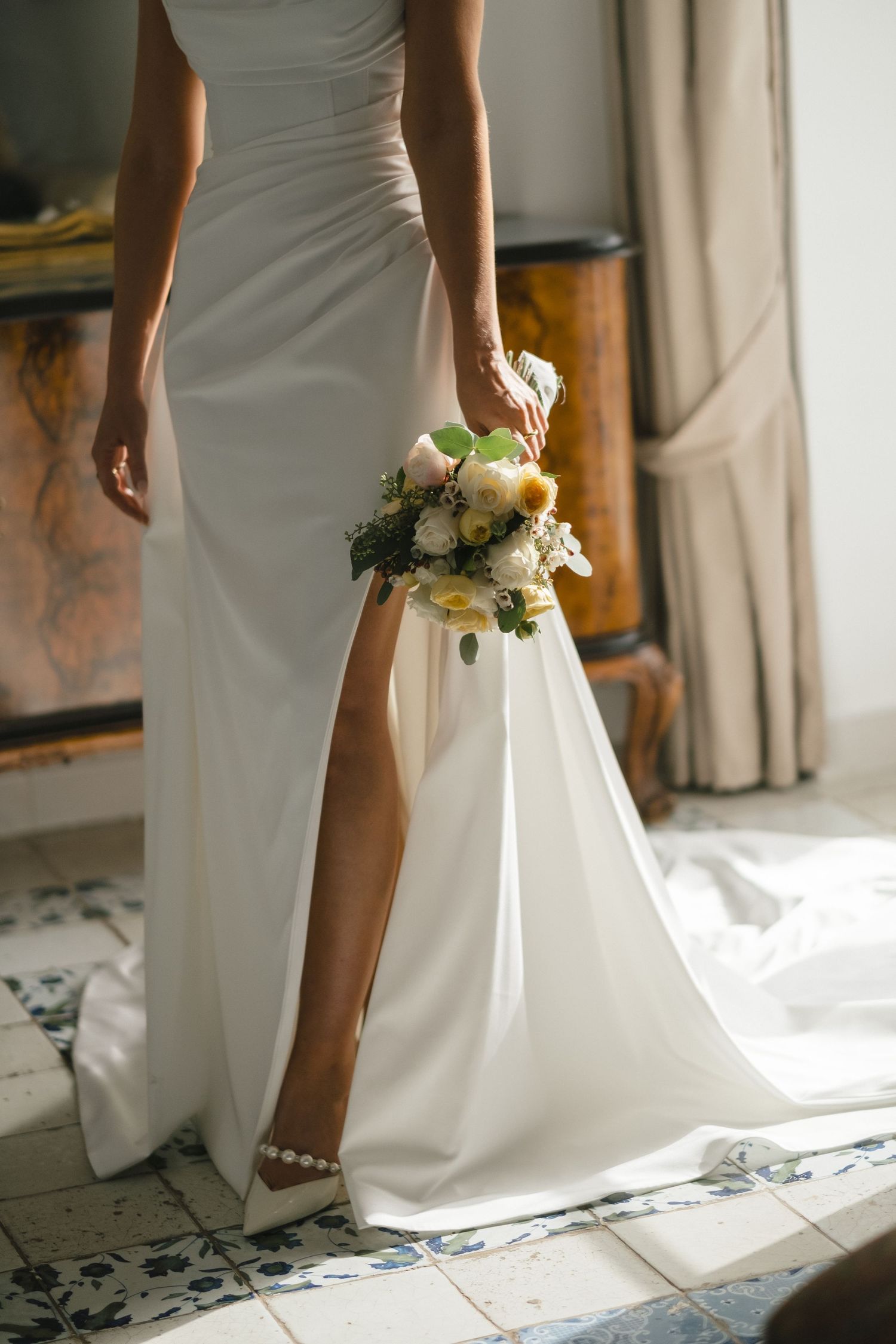 Bride in white slit gown holding a bouquet of white and green flowers, standing on blue tile floor.