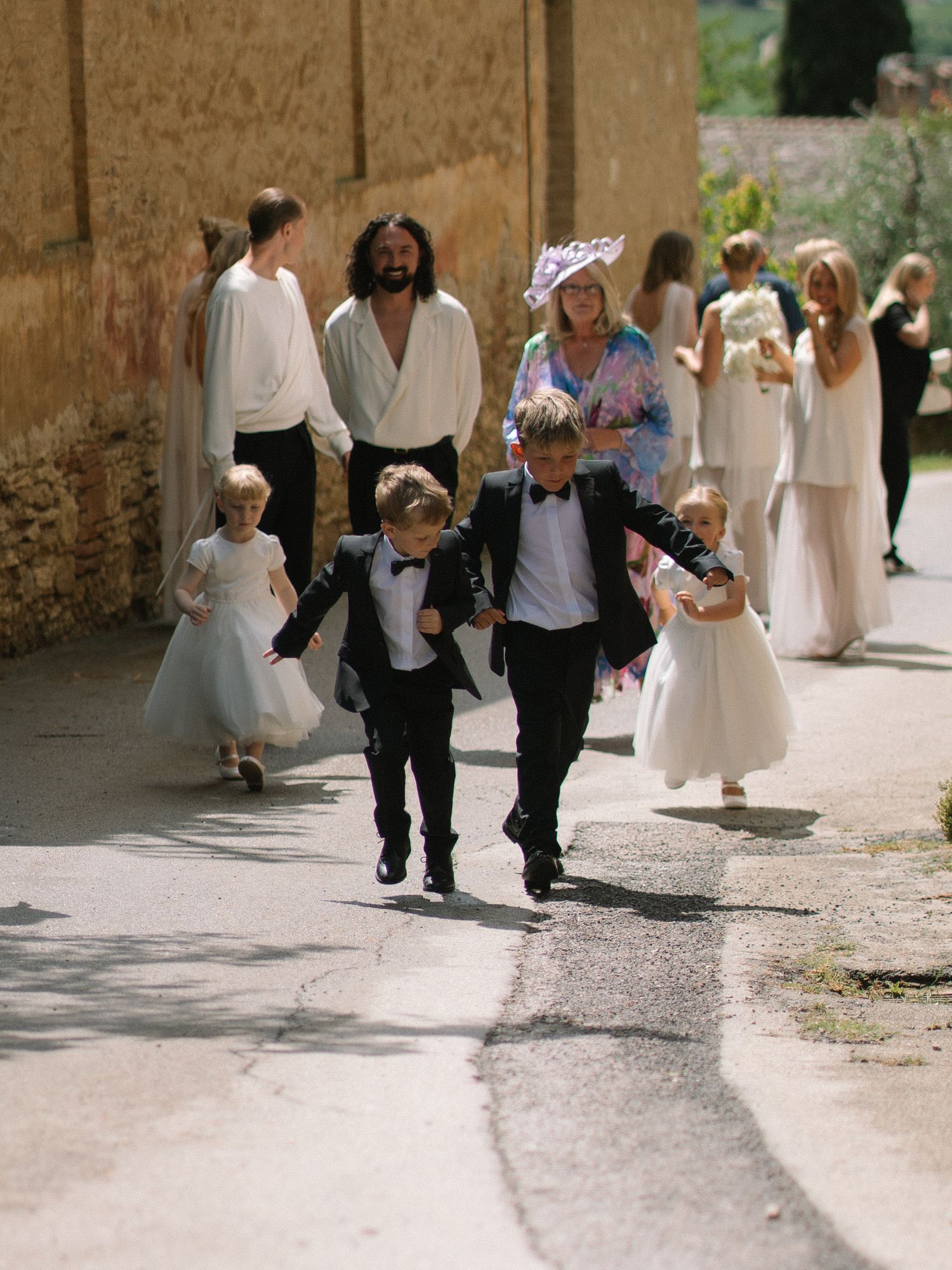 Wedding party processional with flower girls in white dresses and ring bearers in tuxedos walking along a sunlit stone path.