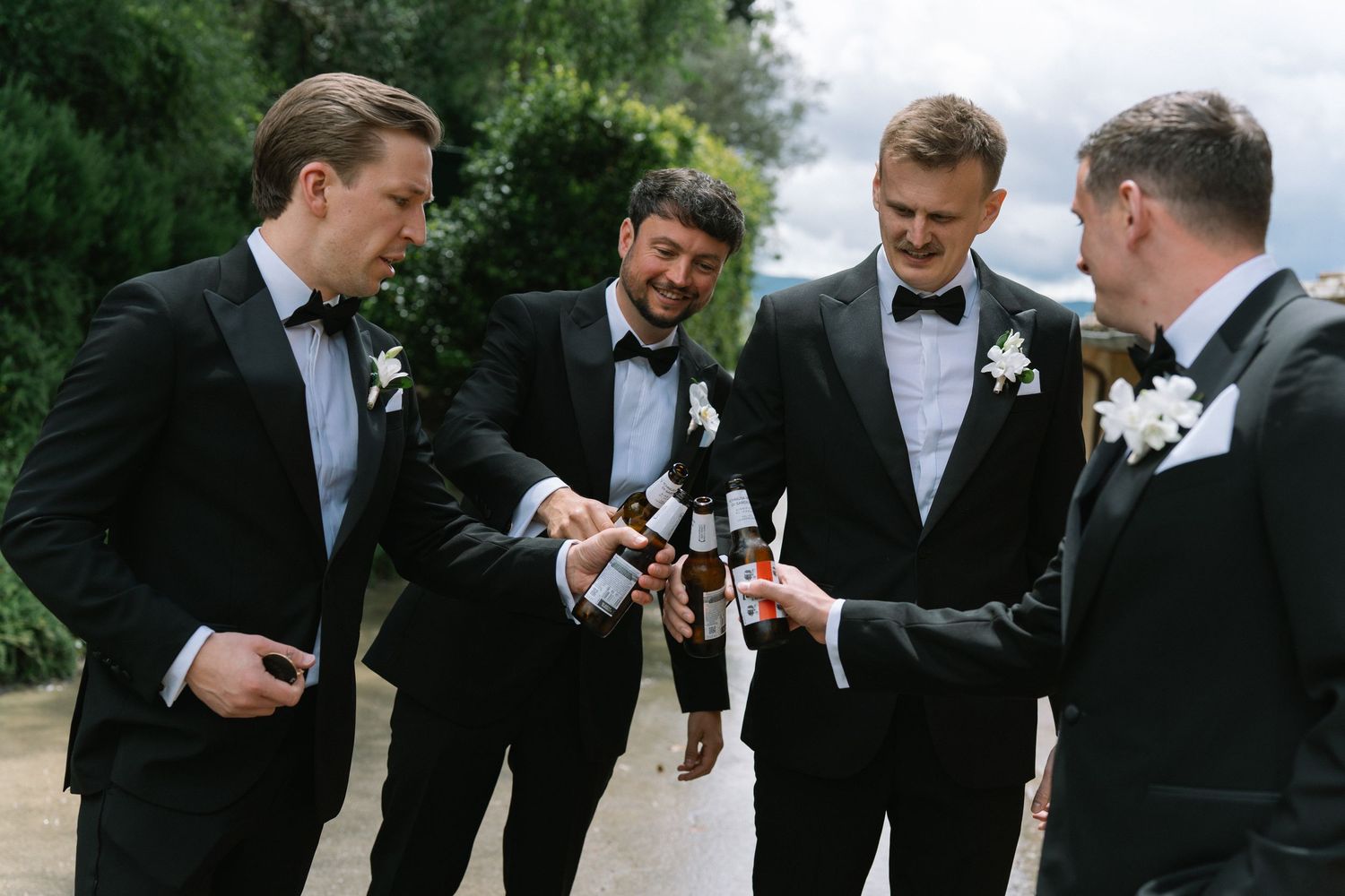Four groomsmen in black tuxedos toasting with beer bottles outdoors at Tuscan wedding venue.
